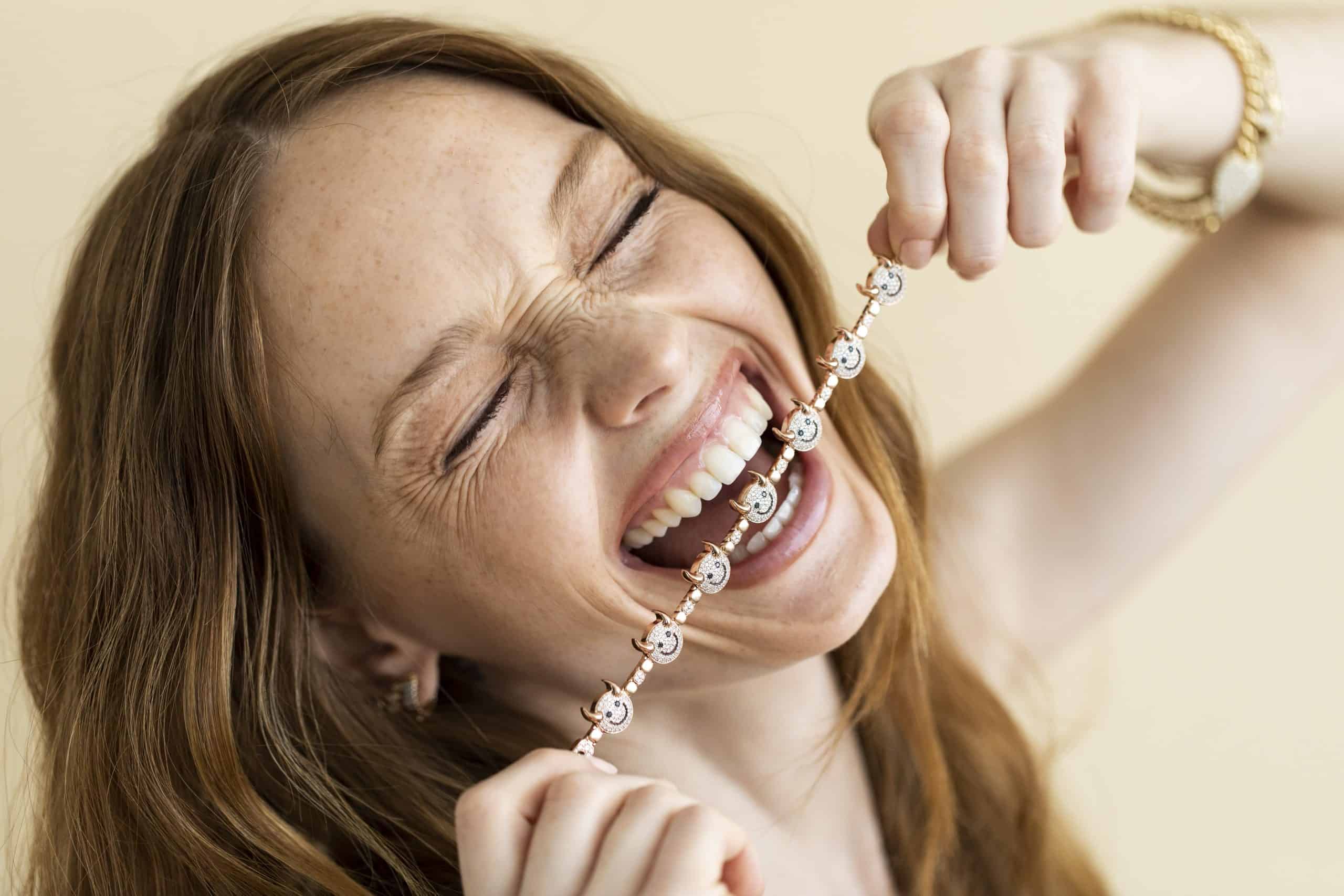 A woman playfully bites her Lucky Devil Bracelet, exuding good fortune. With closed eyes and a beaming smile, she accessorizes elegantly with customizations like a watch and earrings against a soft beige background.