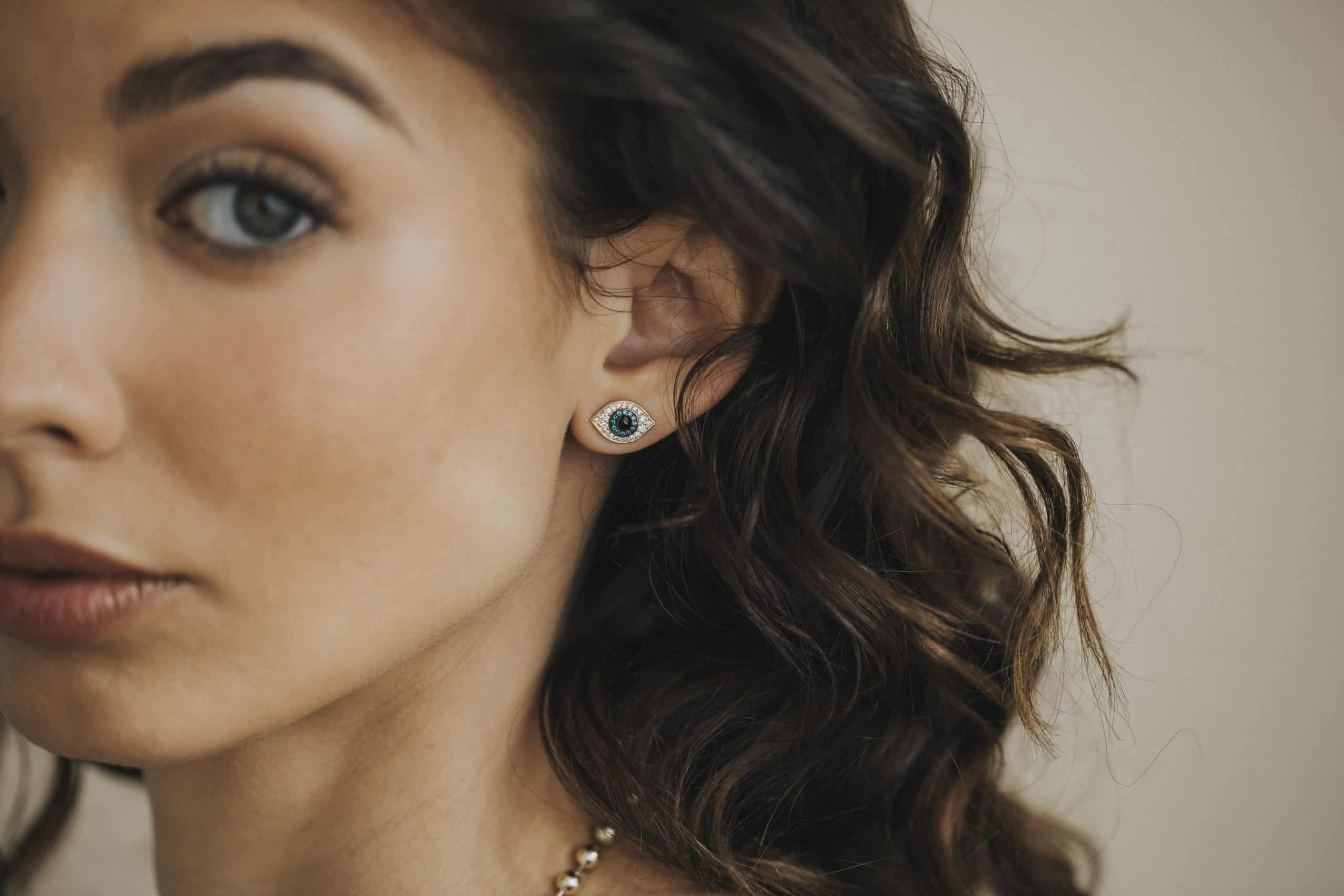 A person with curly brown hair and arched eyebrows wears Janet Heller Fine Jewelrys Diamond Evil Eye Studs, a unique design. The blurred background highlights the artistry of this custom product as they maintain a neutral expression.