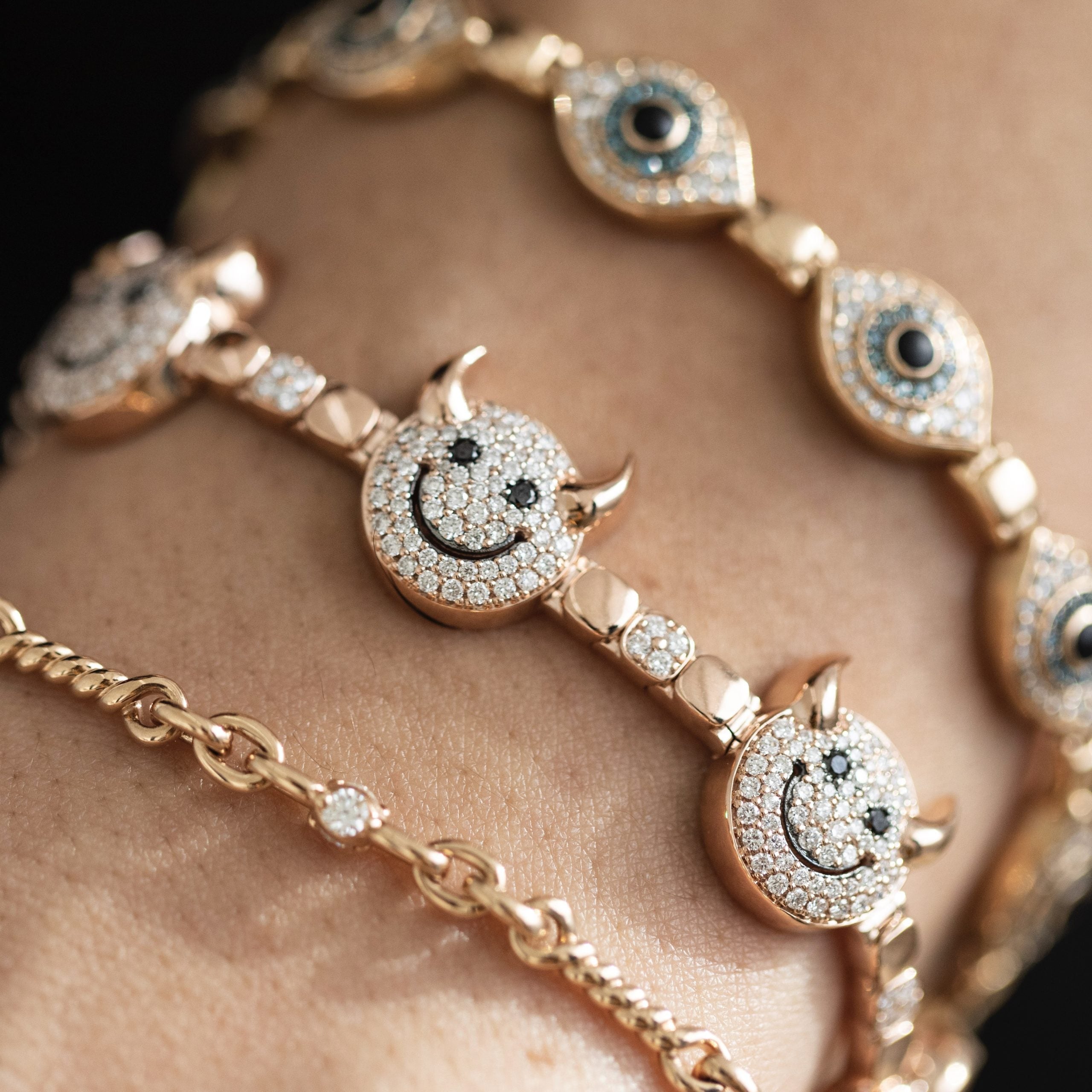A trio of rose gold bracelets adorns the wrist: the Lucky Devil bracelet, featuring devil-horned smiley faces with diamonds; another with eye-shaped charms and dark centers; and the third, a simple classic chain for good fortune.
