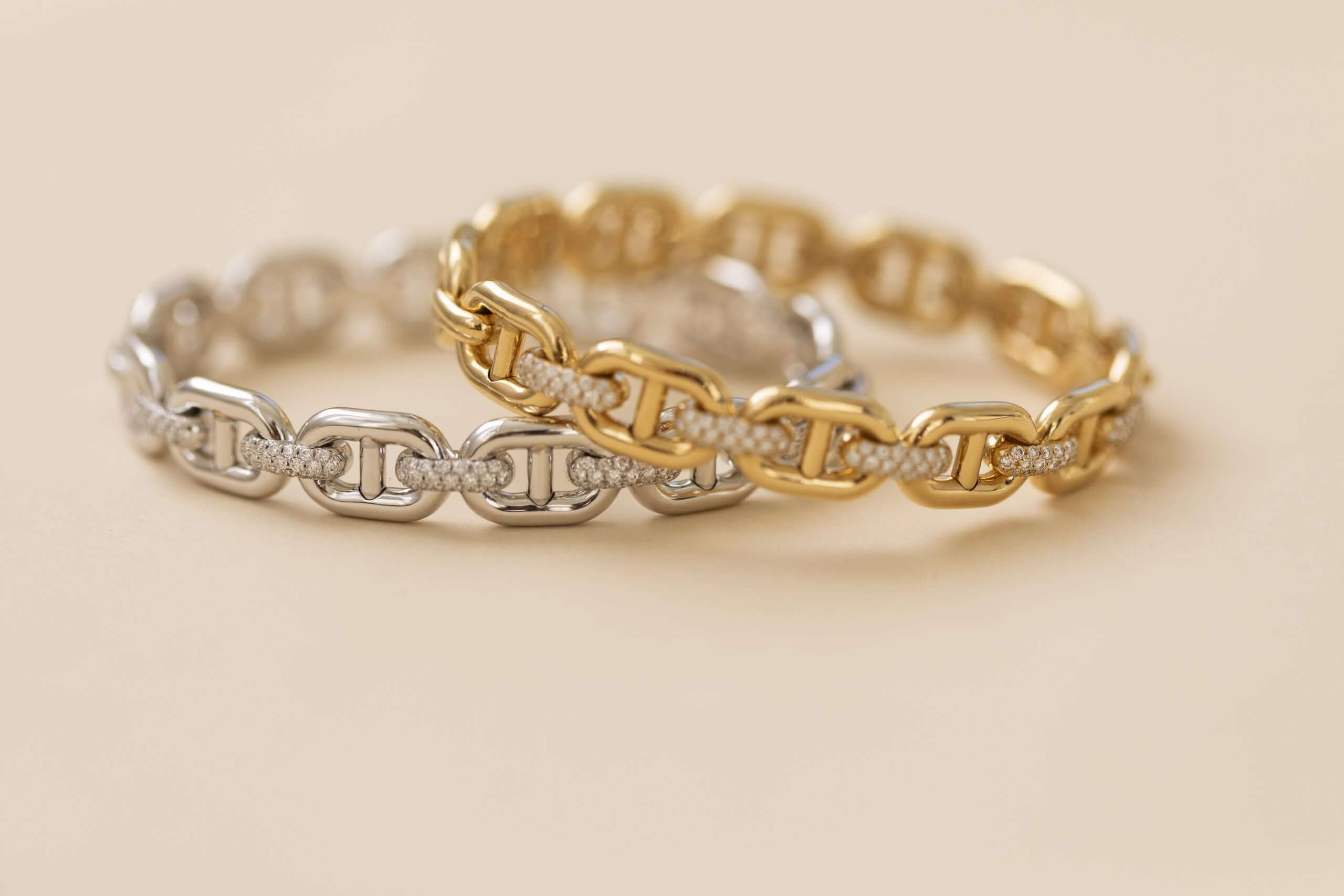 Two Diamond Mariner Link Bangles from Janet Heller Fine Jewelry—one in silver tone and one in gold tone—are displayed side by side on a light beige surface. Each bangle features an elegant, polished finish with sparkling diamond accents on select links, exemplifying the brand's signature style. For jewelry inquiries or custom requests, please reach out directly.
