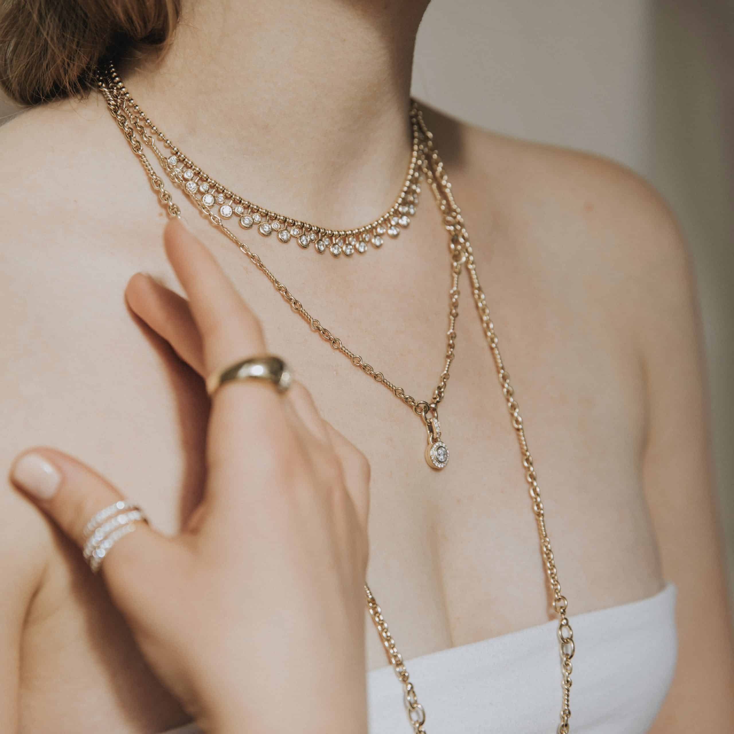 The person elegantly showcases fine jewelry, wearing layered gold necklaces and the Diamond Alexander Necklace. A gold ring graces their index finger, while a silver ring with small gems adorns their pinky, all beautifully complemented by janethellerfinejewelry against a white strapless top.