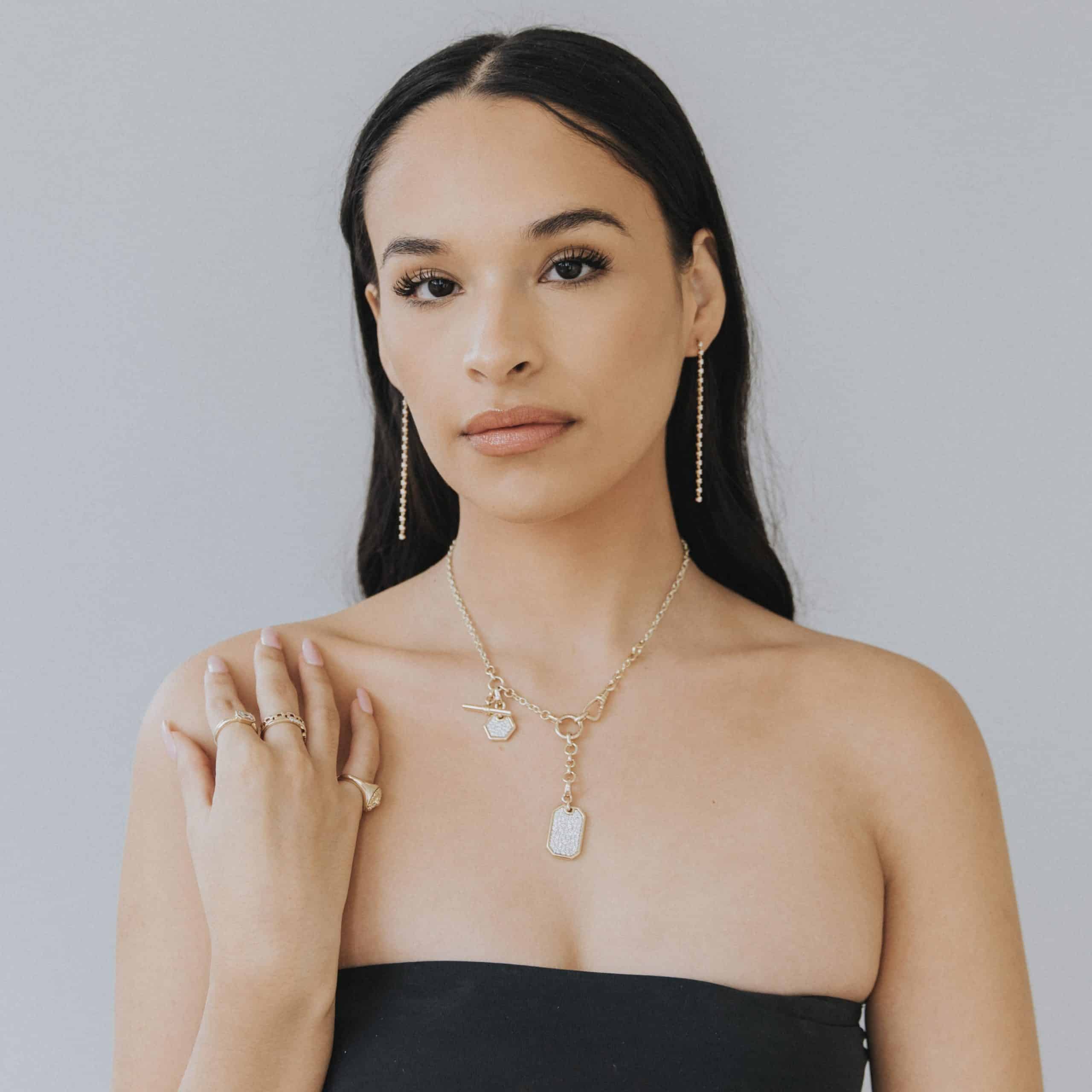 A woman with long dark hair and a neutral expression wears a strapless black top. She showcases Janet Heller Fine Jewelrys Berlin Chain necklace with charms, matching dangle earrings, and several rings. Her hand gently rests near her collarbone against the light gray background.