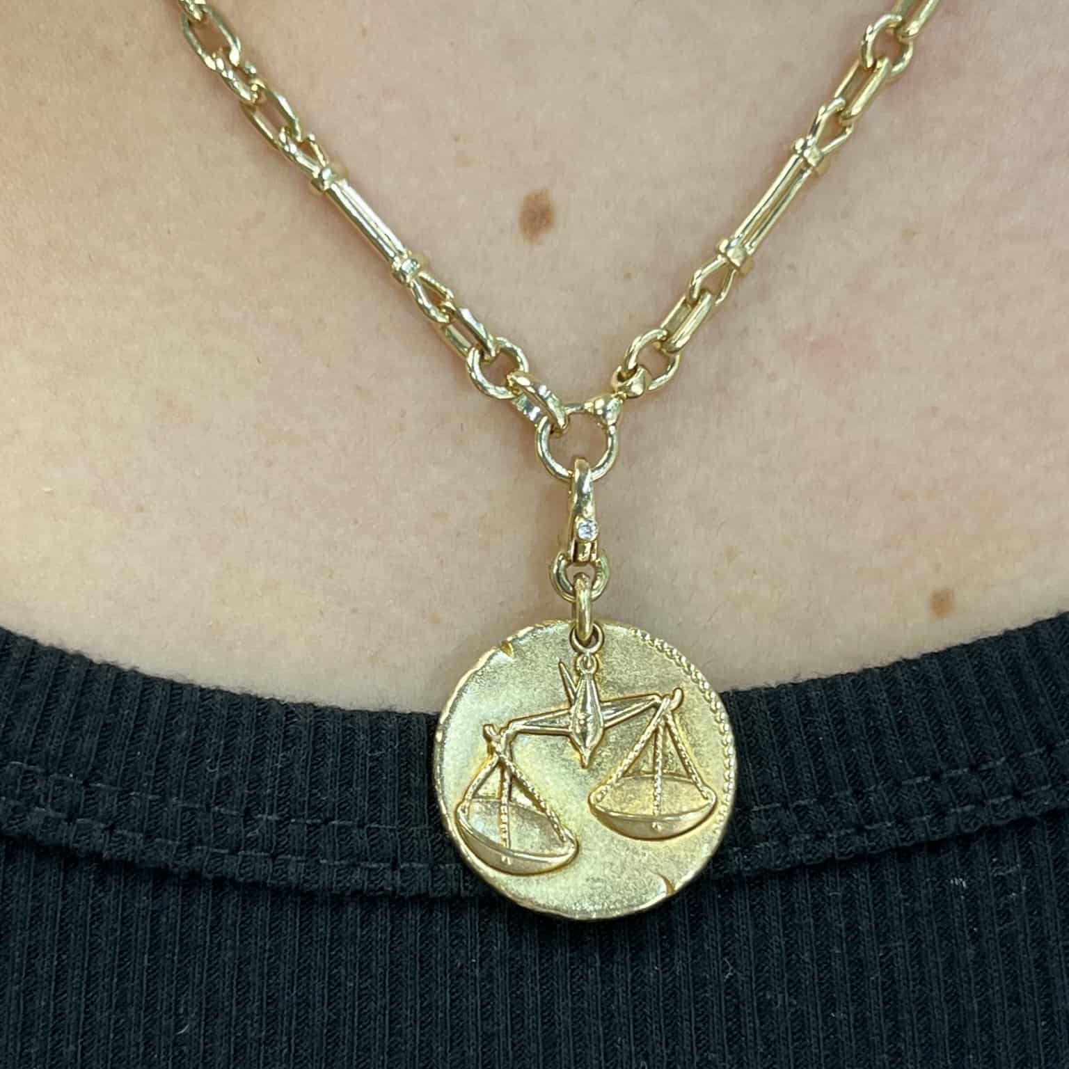 Close-up of a gold Zodiac Sign Charm necklace with a clasp, featuring a justice scales pendant. The linked chain and textured pendant rest elegantly against a black fabric background.