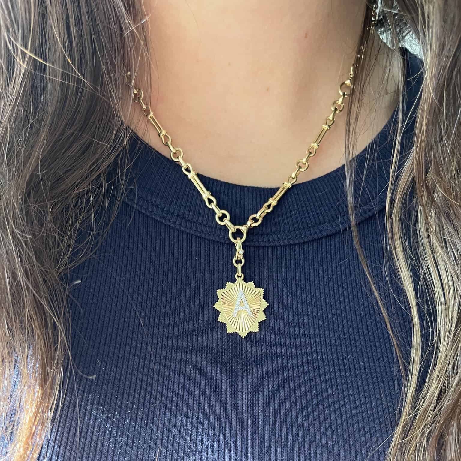 A person models a Janet Heller Fine Jewelry piece: the Lisbon Chain, a gold necklace, over a navy blue ribbed top. The chain-link design beautifully complements the intricate sunburst pendant as long brown hair partially frames this charming accessory.