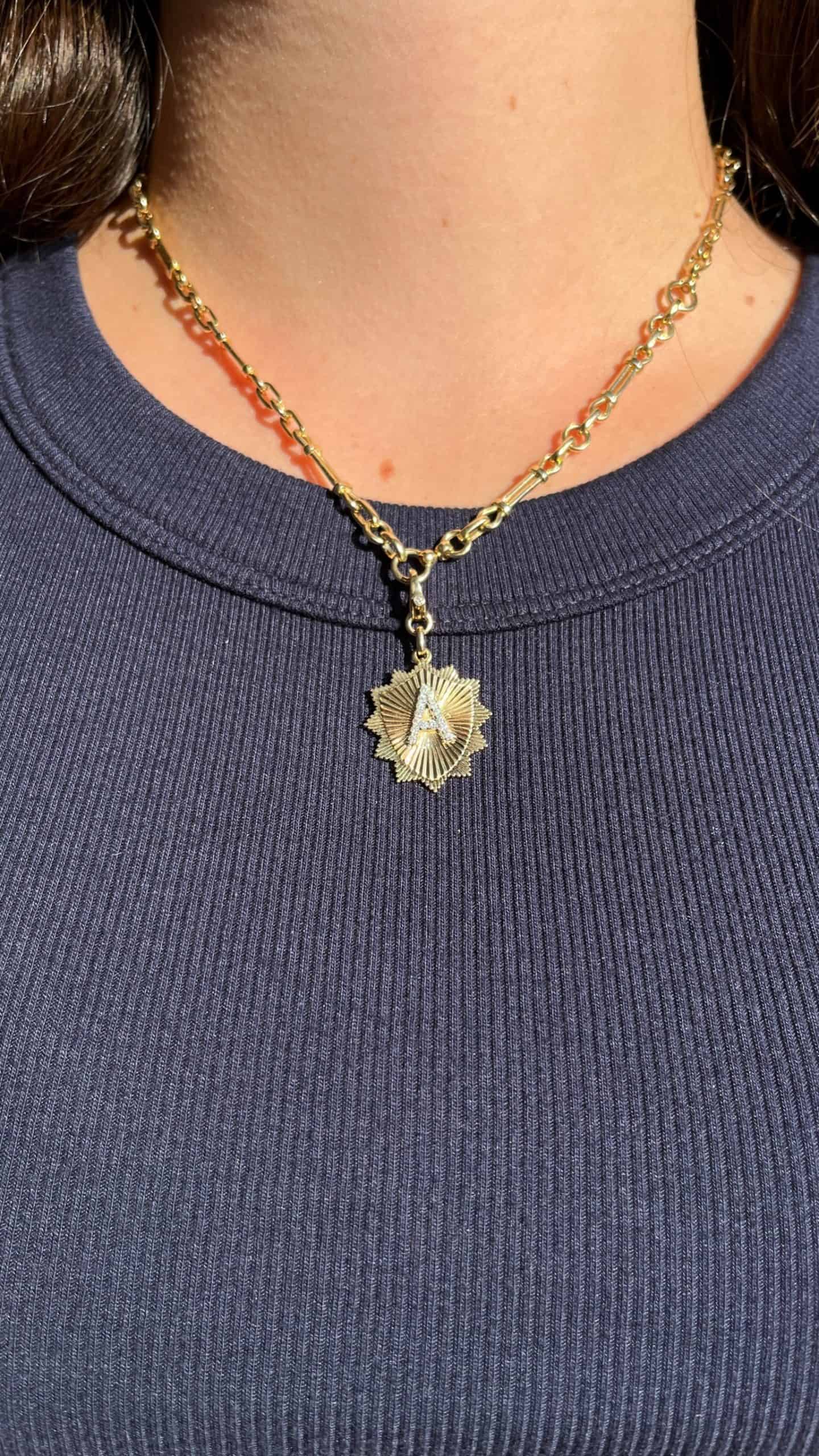 A person wearing a ribbed navy top and a gold chain necklace with the Diamond Initial Shiloh Charm by Janet Heller Fine Jewelry catches the sunlight. The intricate detailing and elegant clasp make it both captivating and secure.