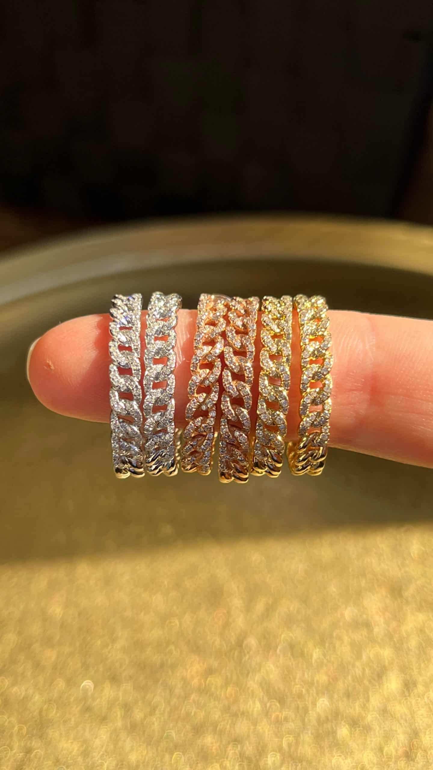 A hand displays the Chain Link Easton Hoops by Janet Heller, featuring five sparkling hoop earrings in silver, rose gold, and gold tones, each encrusted with small diamonds, set against a blurred golden background—an exquisite showcase of fine jewelry.
