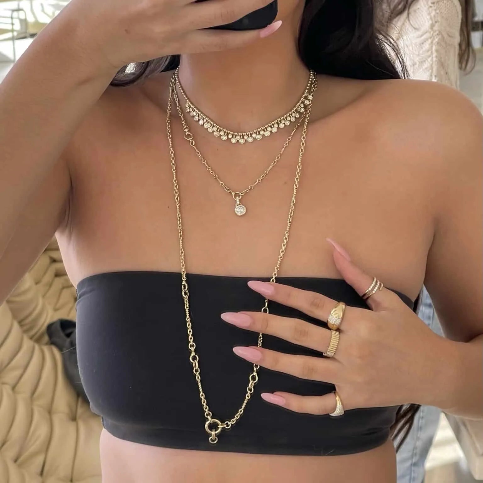 Wearing a black tube top, a person poses for a selfie featuring fine jewelry, including the Diamond Alexander Necklace and other pieces from janethellerfinejewelry. Their long nails are light pink, and they hold a smartphone that partially covers their face.