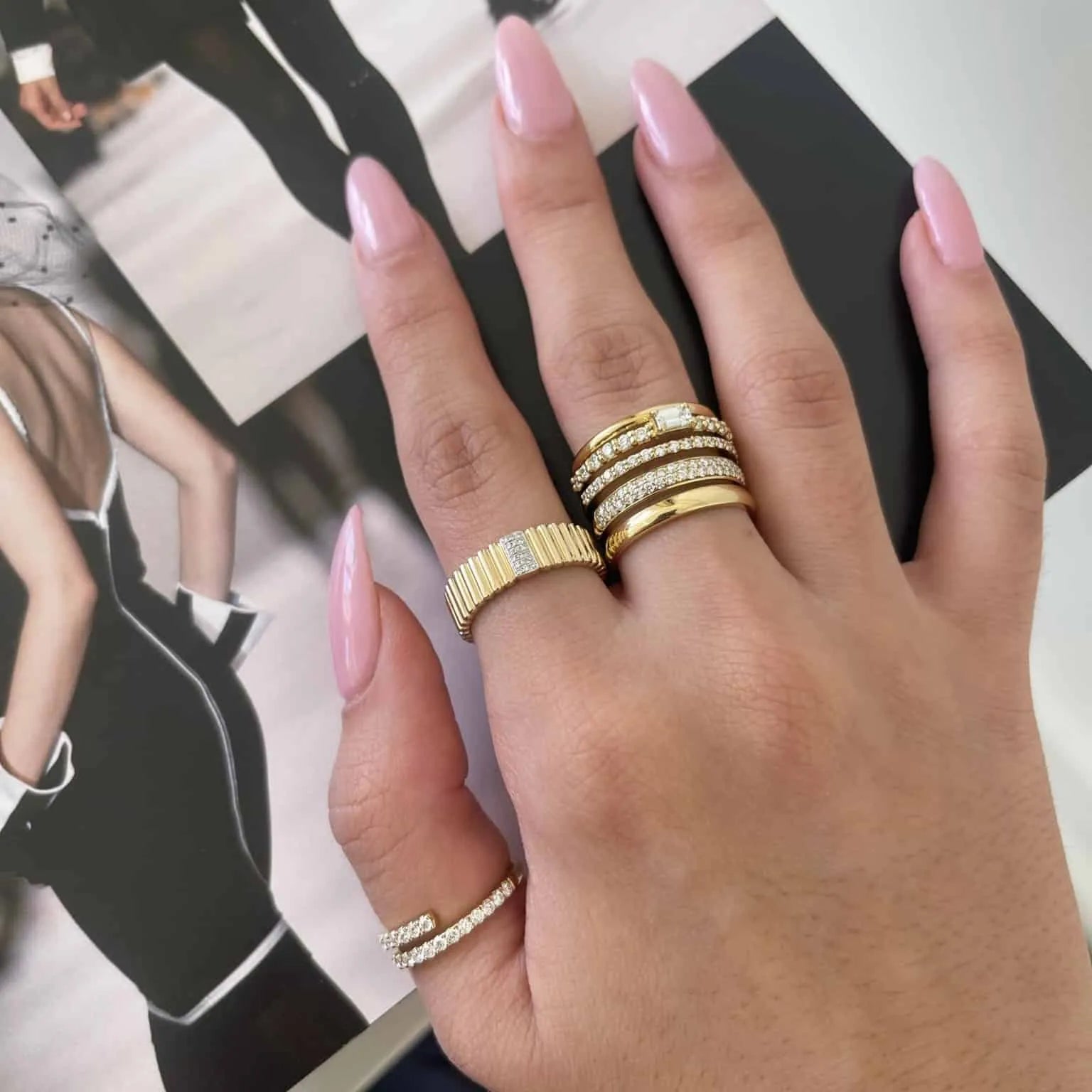 A hand with long, polished pink nails, adorned with fine jewelry including the Diamond Sloane Ring, rests on an open fashion magazine featuring a chic black outfit.