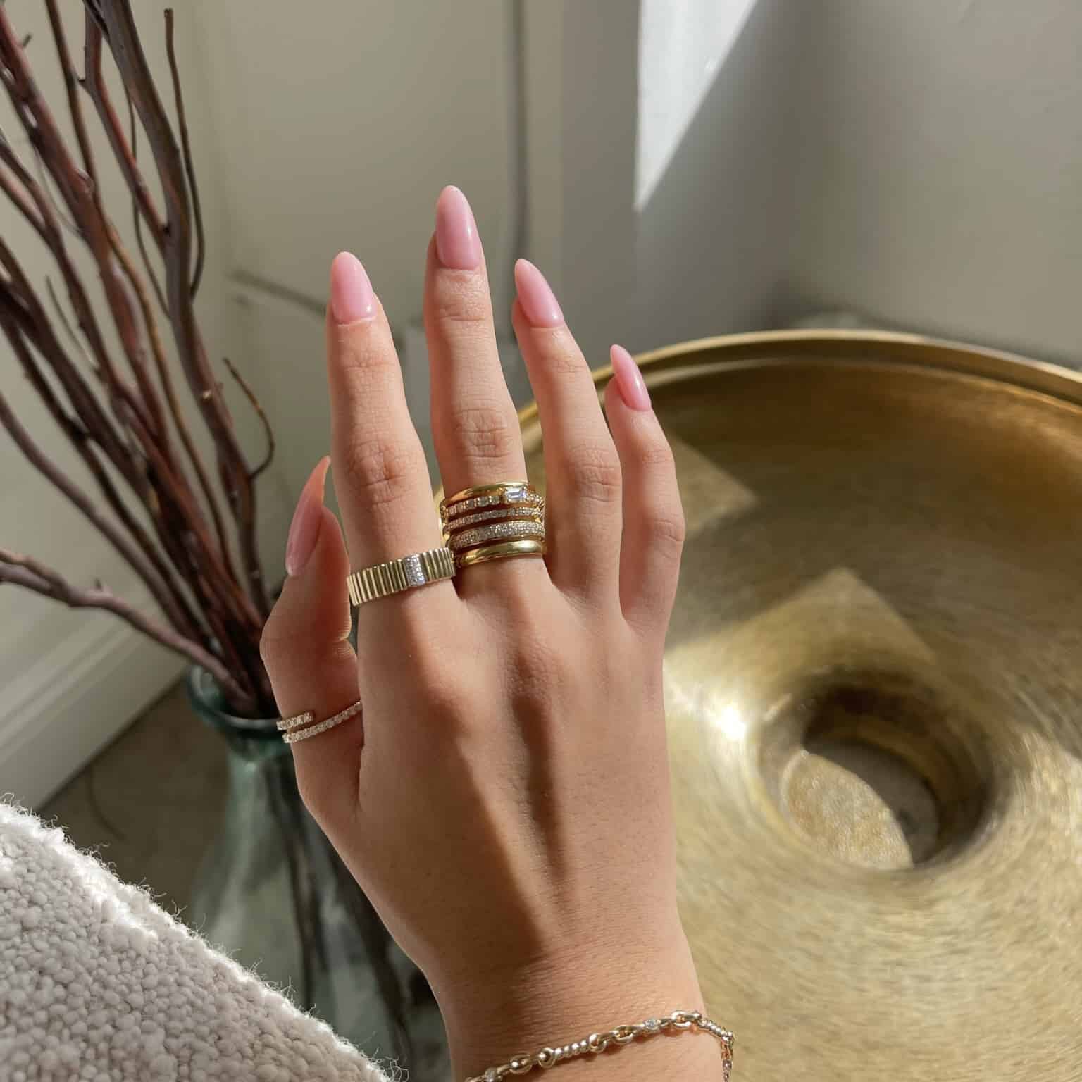A hand with long, pink nails displays elegant pieces of jewelry adorned with the Diamond Sloane Ring. The backdrop includes a metallic decorative dish and tall dried branches. The person dons a cream-textured sweater. Preorder the Diamond Sloane Ring now to elevate your style.