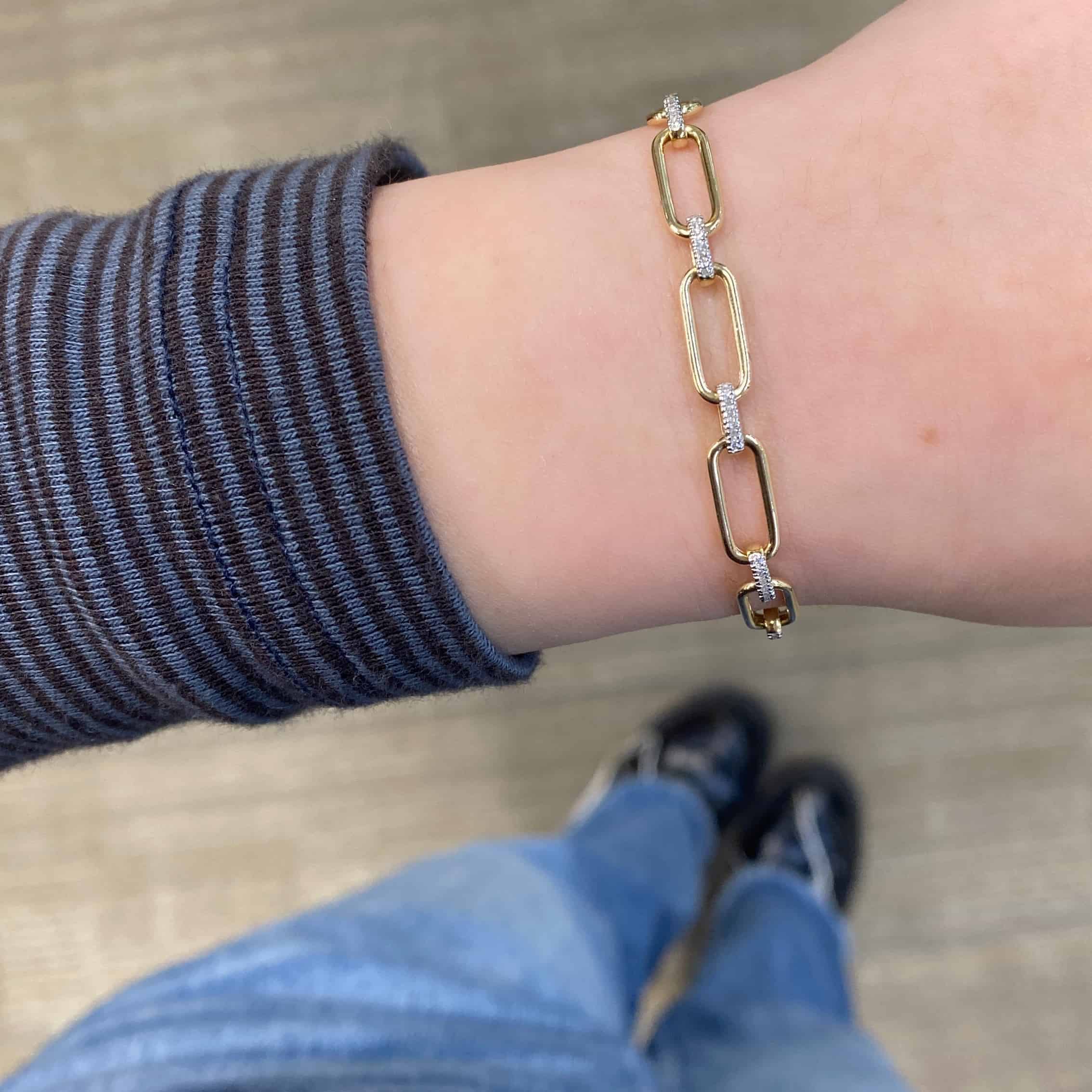A close-up of a wrist adorned with the Diamond Oval Link Bracelet, featuring gold links and small diamonds. The individual wears a striped long-sleeve shirt, blue jeans, and black shoes over a light brown textured floor.