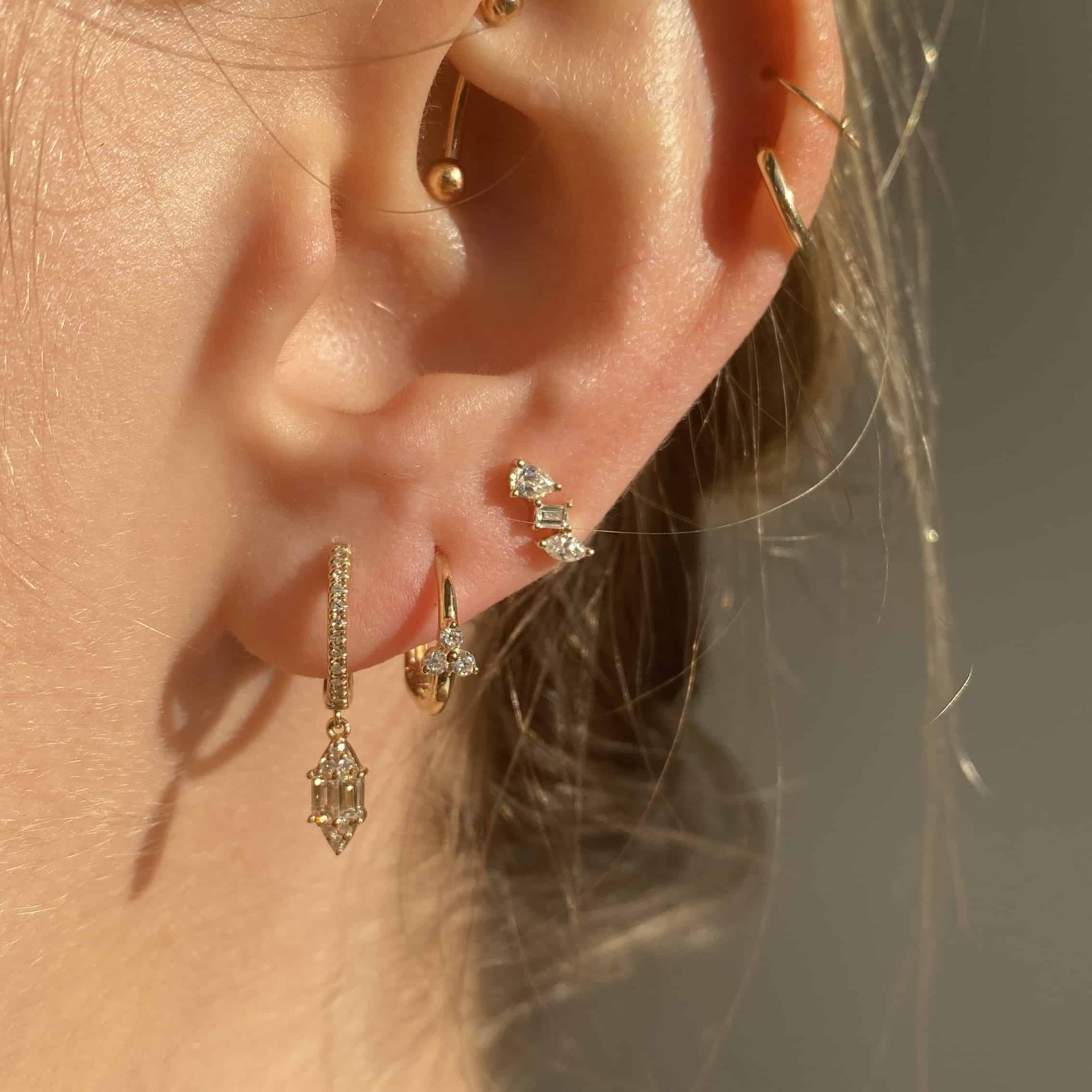 Close-up of an ear featuring Janet Heller Fine Jewelry, showcasing four exquisite piercings: Single Diamond Cluster Huggies, a small hoop with a geometric charm, a stud with three stones, and two customizations—a hoop with a clear stone and a simple upper-ear hoop.