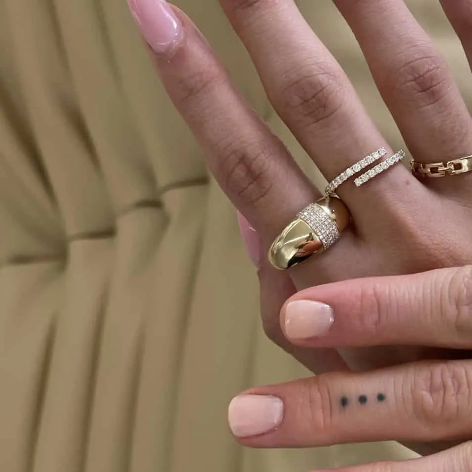 Close-up of a hand with nude manicured nails. The index and middle fingers sport fine gold jewelry; one ring features an Extra Large Diamond Dome design. A tattoo of three dots is visible on the fingers side against a beige fabric background.