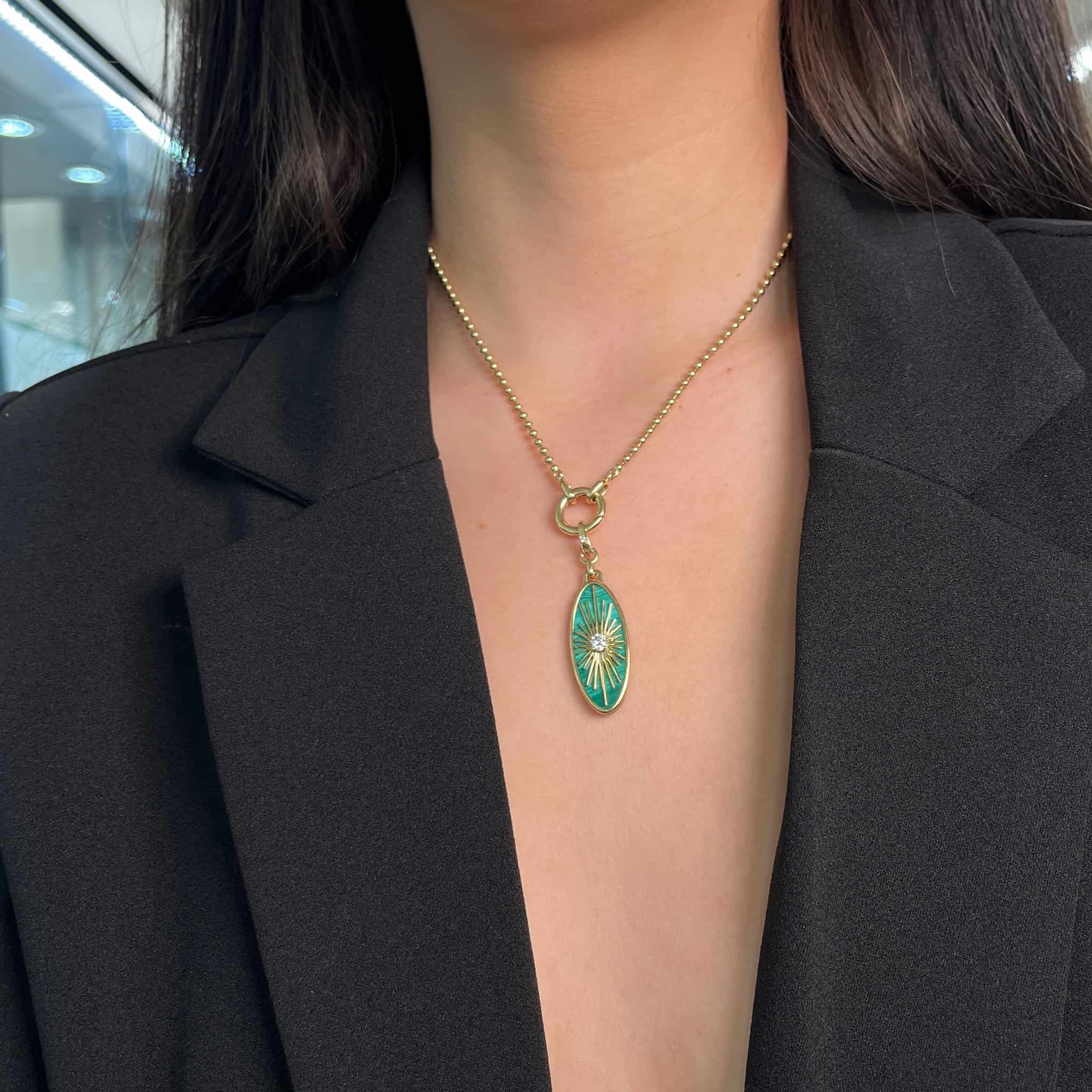 Wearing a black jacket with a deep neckline, a person showcases the Malachite & Diamond Oval Charm from Janet Heller Fine Jewelry. The gold necklace features an oval green pendant with a starburst design, beautifully highlighted against a softly blurred background.