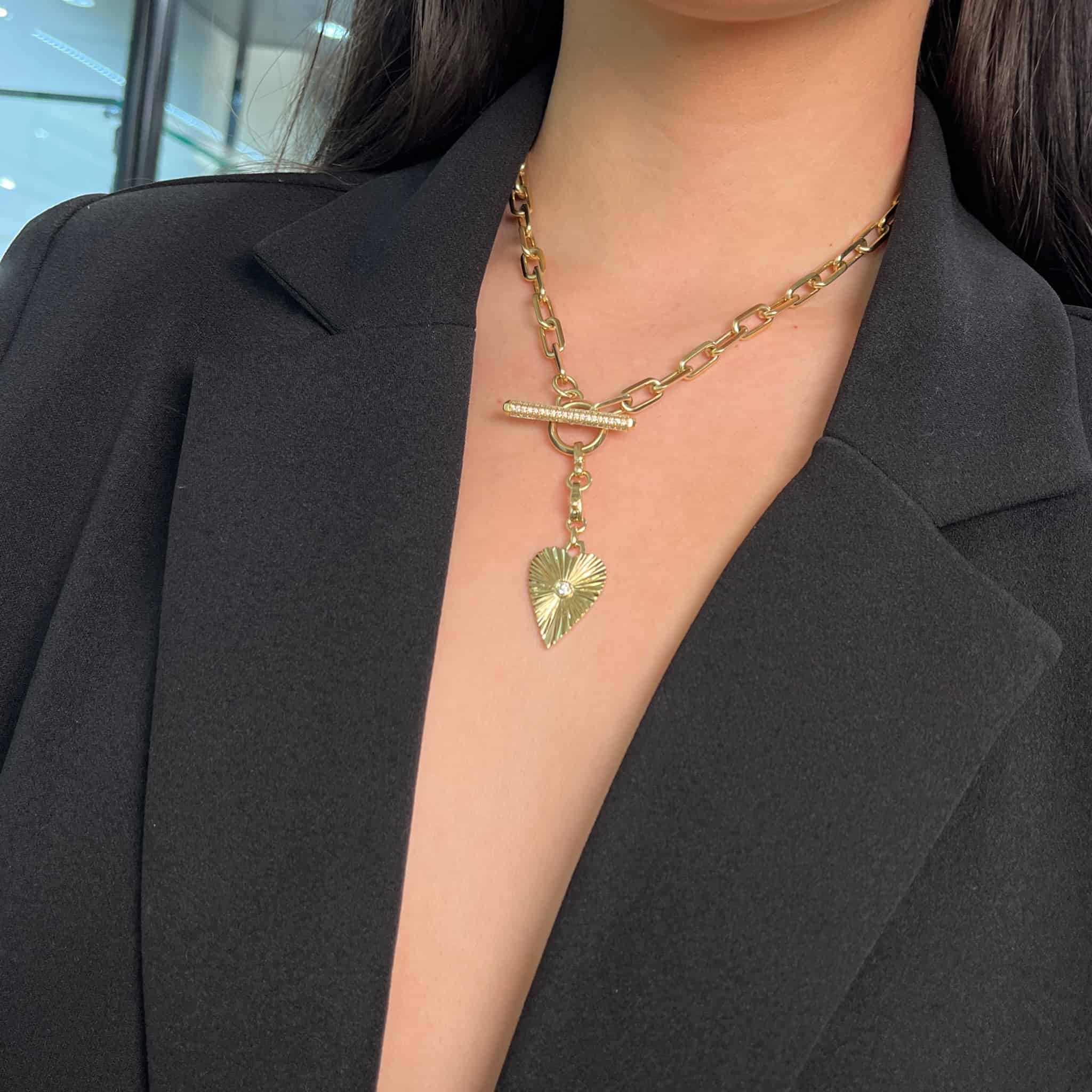 Close-up of a person wearing a black blazer with a Janet Heller Fine Jewelry gold chain necklace. The Diamond Heart Burst Charm, featuring a heart-shaped pendant and toggle clasp, is customizable. A blurred modern interior serves as the background.
