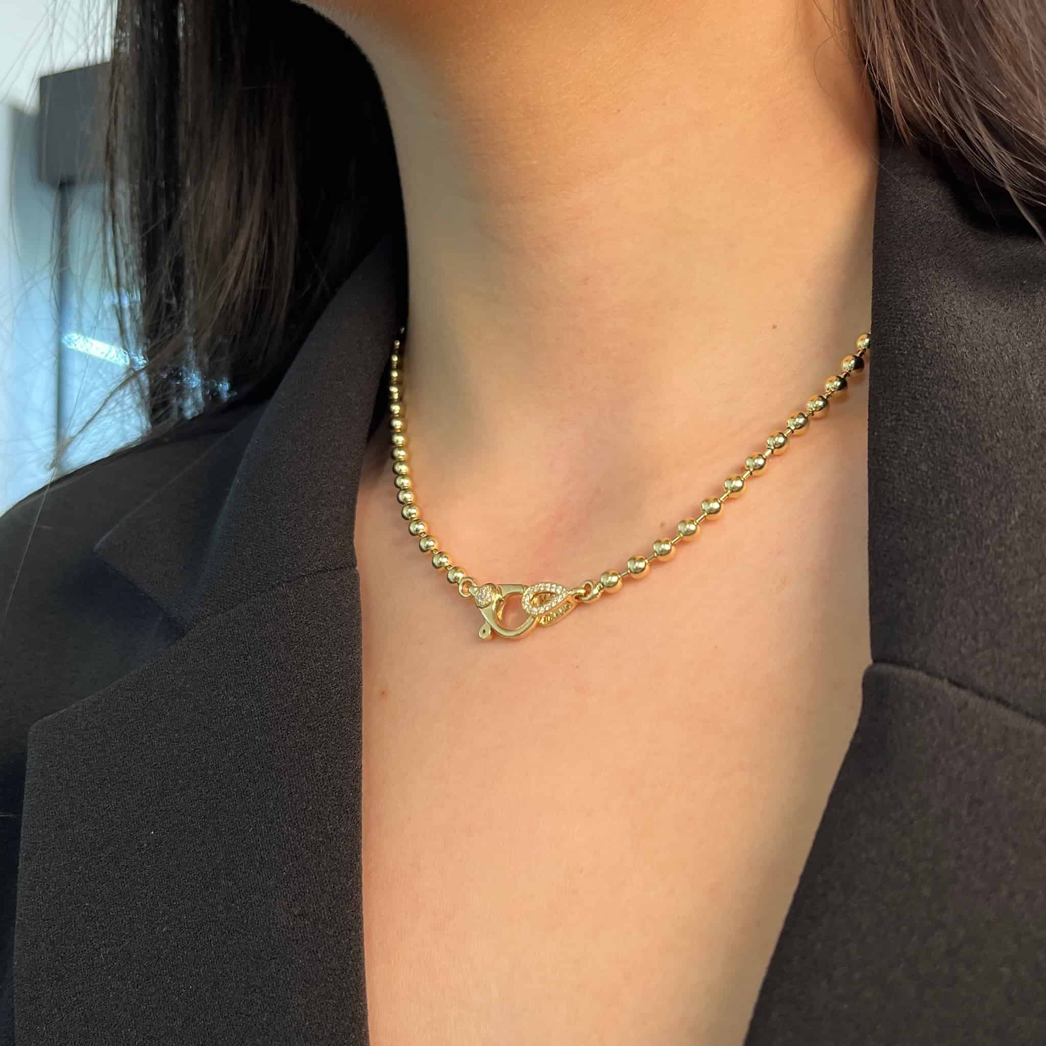 A person in a black blazer wears Janet Heller Fine Jewelrys 4mm Paris Chain, featuring a gold beaded necklace with a decorative clasp. The close-up highlights the exquisite jewelry and attire around the neck and collar area.
