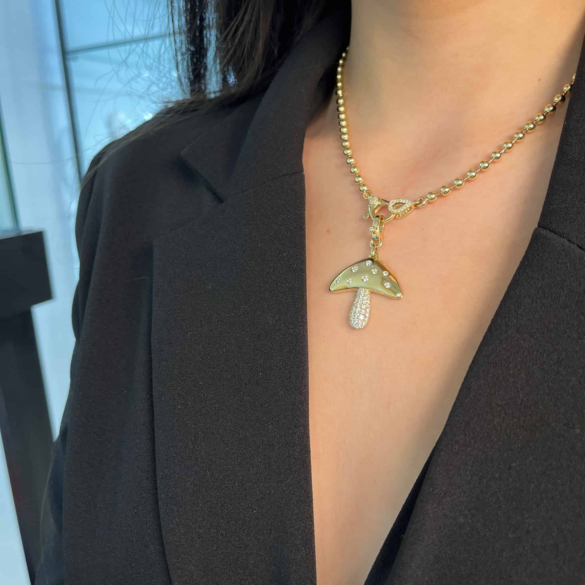 A close-up shows a person in a black blazer wearing Janet Heller Fine Jewelrys Diamond Mushroom Charm, featuring mushroom-shaped charms with small gems. The expertly crafted pendant clasp is highlighted against the backdrop of a softly lit indoor window.