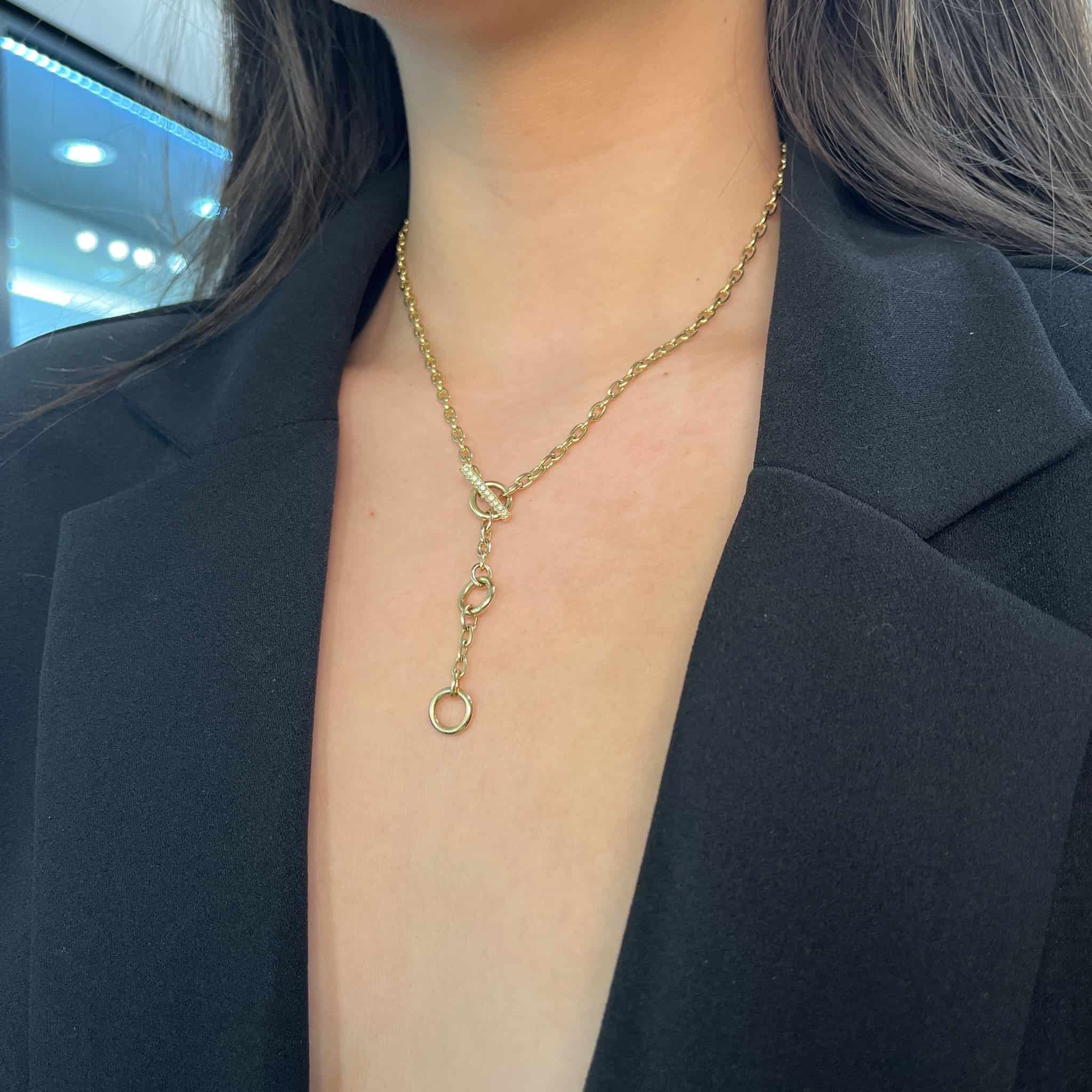 A person in a black blazer with an open neckline wears the Janet Heller Fine Jewelry London Chain gold necklace, adorned with two circular charms. The background is softly blurred with blue and white lights.