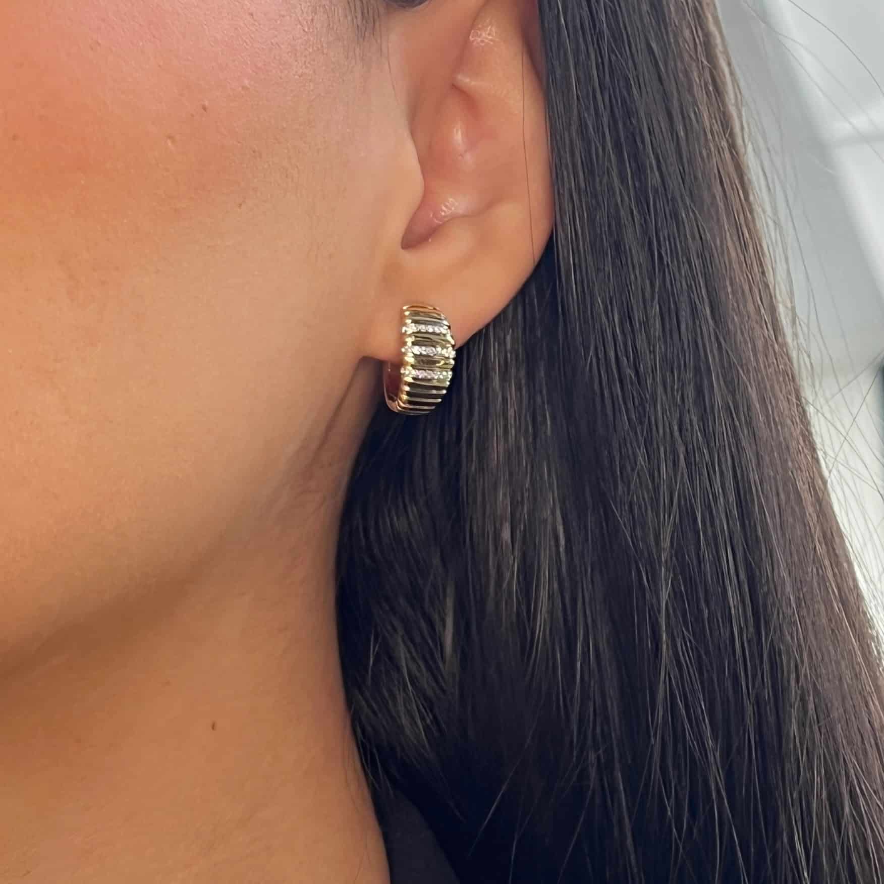 Close-up of someone wearing Diamond Ridge Earrings, small gold hoops with a ribbed design from janethellerfinejewelry. Their ear and straight dark hair stand out against a blurred backdrop, highlighting exquisite craftsmanship and customization potential.