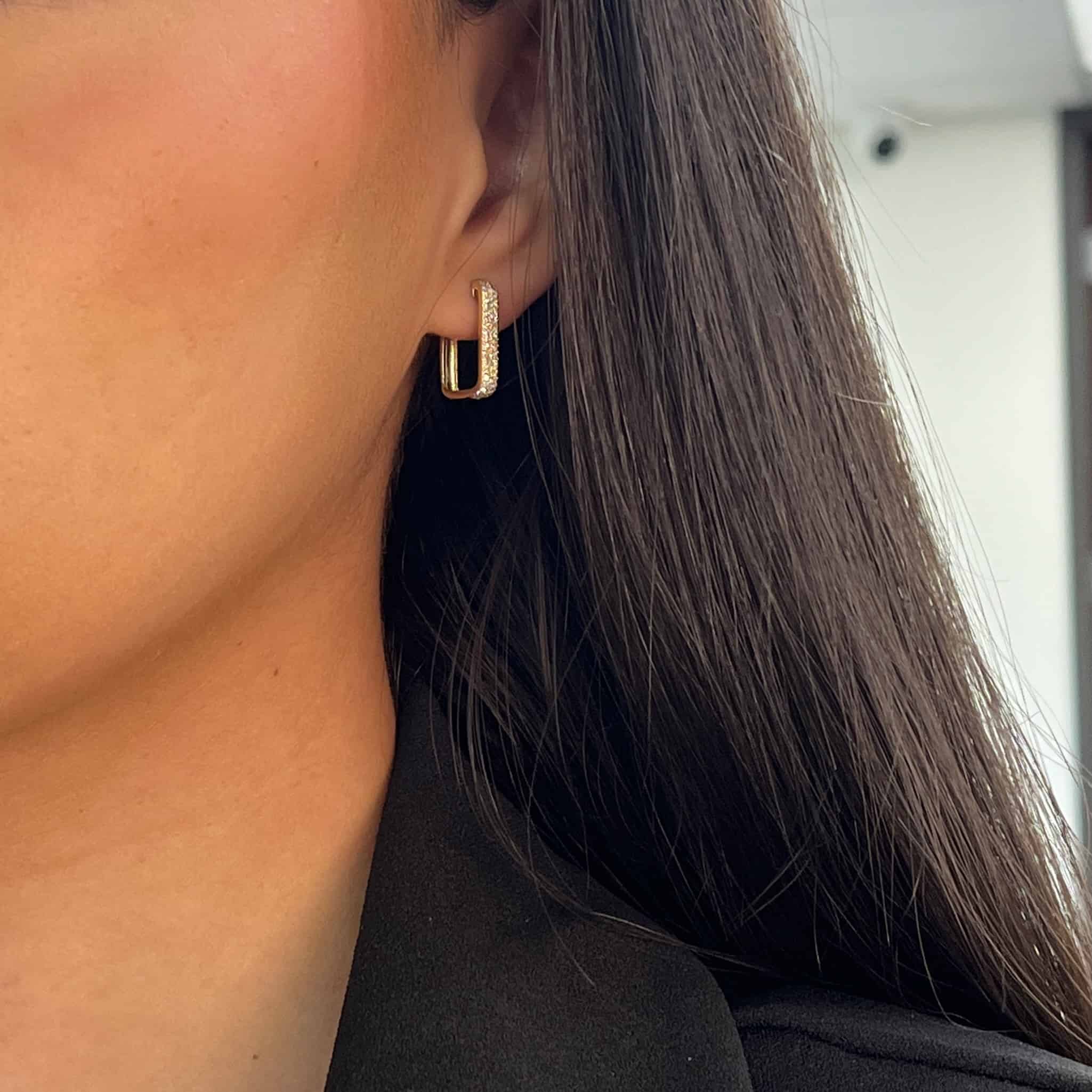 A close-up shows a person with long dark hair wearing Janet Heller Fine Jewelrys Pave Diamond Square Hoops and a black garment, set against a softly blurred background. Contact us to customize your stunning piece.