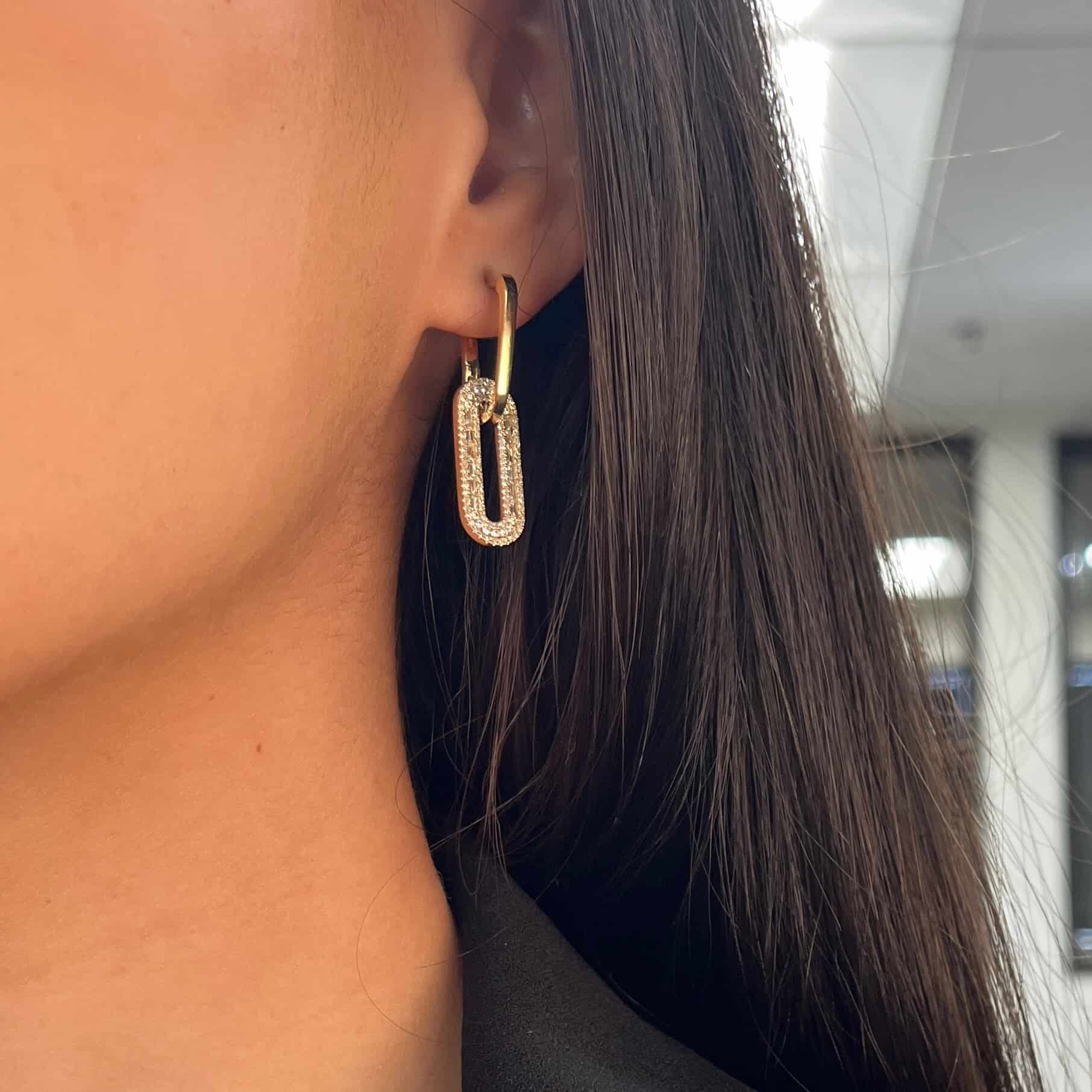 A close-up showcases FoundryFineJewelrys exquisite Baguette Diamond Oval Link Earrings: gold hoops with a hanging rectangular gemstone pendant. The blurred background highlights the intricate details, drawing attention to the product and the persons ear and hair.