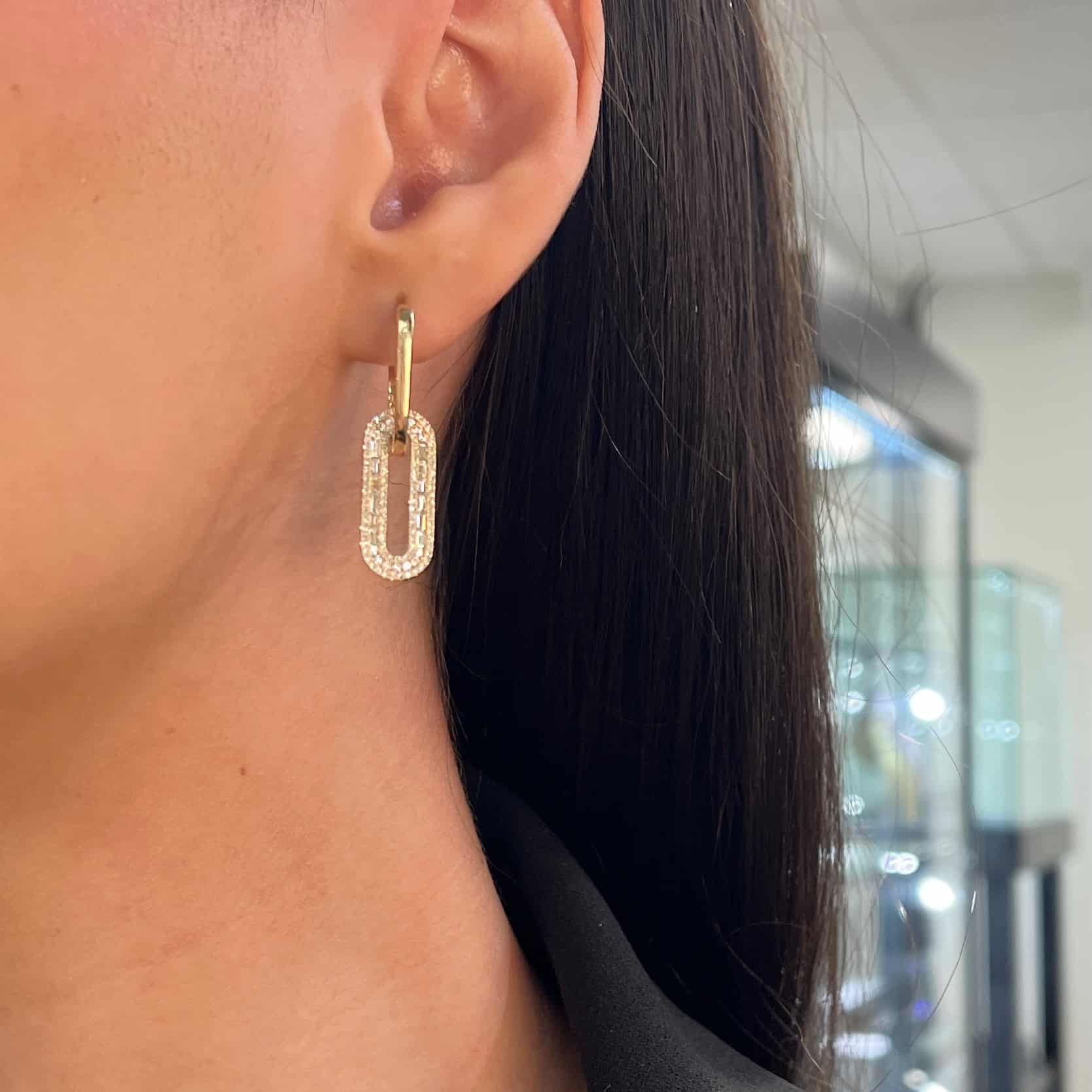 A person with long dark hair wears FoundryFineJewelrys Baguette Diamond Oval Link Earrings—a stunning piece of jewelry. The softly lit room and glass display enhance the products elegance.