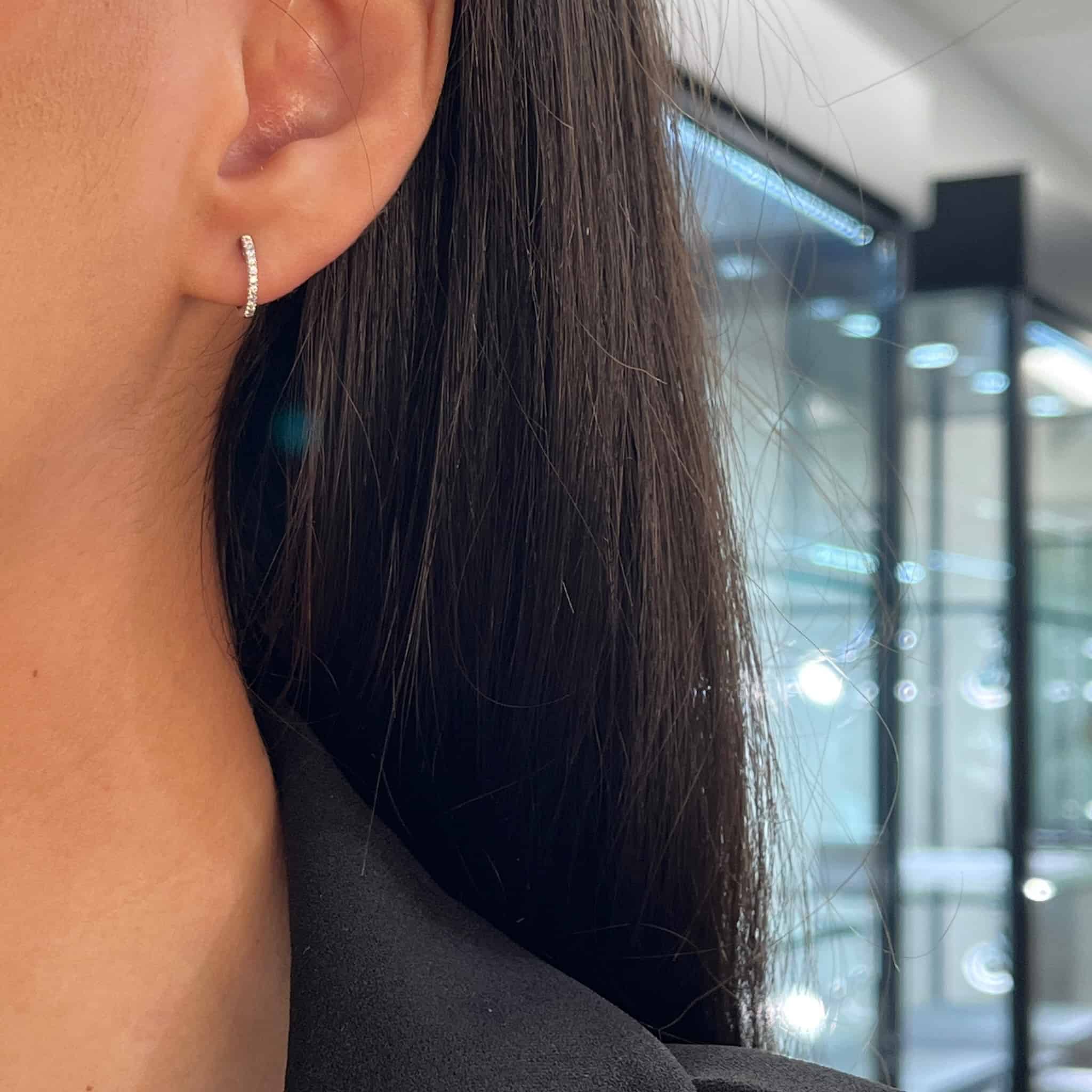 A close-up of Janet Hellers ear highlights the FoundryFineJewelry Diamond Marley Huggies, a small, curved earring adorned with sparkling stones. Her long, dark hair elegantly drapes over a black top against the backdrop of a modern, brightly lit interior with gleaming glass displays.