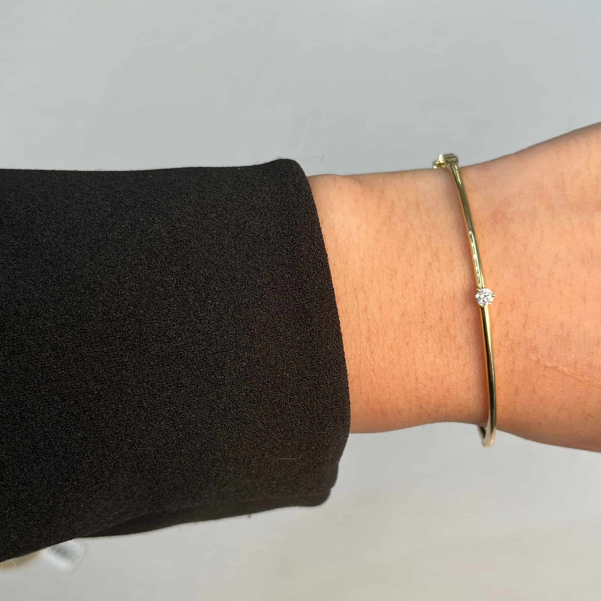 A wrist wrapped in a black sleeve and a delicate gold Diamond Solitaire Bangle with a small diamond from FoundryFineJewelry radiates elegance, standing out beautifully against the plain, light-colored background.