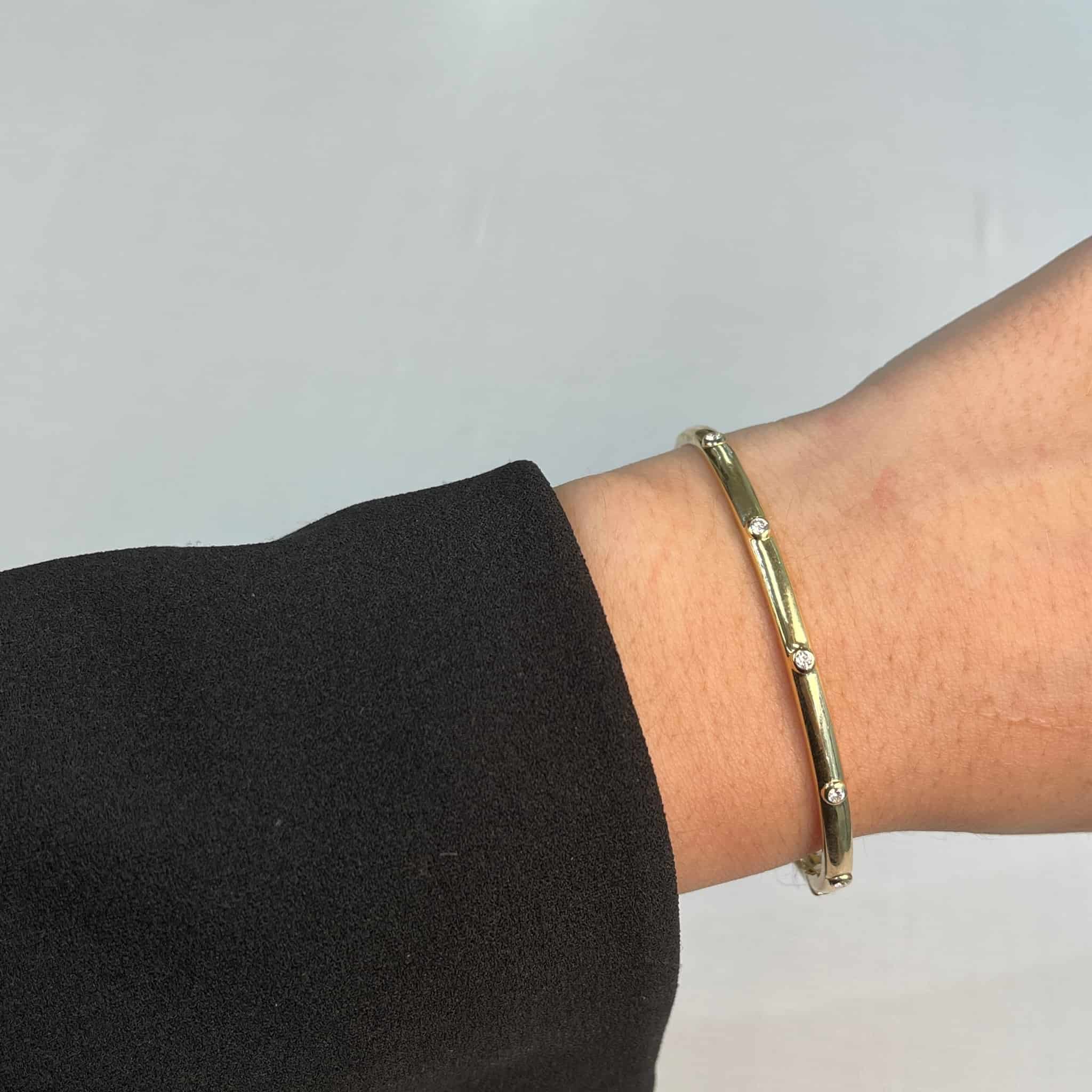 A person wearing a black sleeve showcases the Janet Heller Fine Jewelry Diamond Studded Bangle. Featuring evenly spaced small diamonds against a plain gray background, this elegant gold bracelet invites customizations and inquiries for personalized luxury seekers.