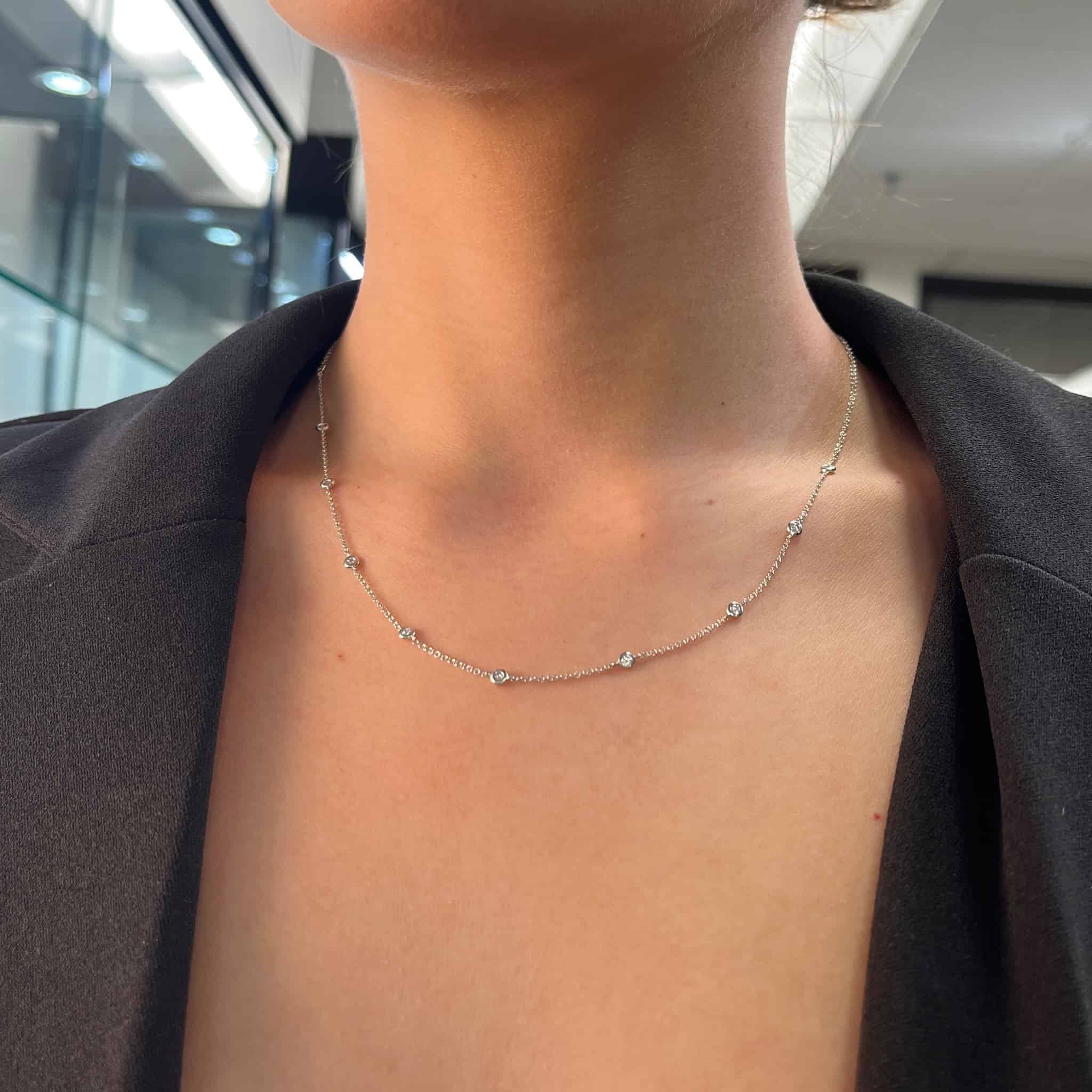 In a blurred indoor setting with lights, a person in a black blazer elegantly displays the Diamonds By The Yard Necklace, a thin silver piece adorned with small, evenly spaced gemstones.