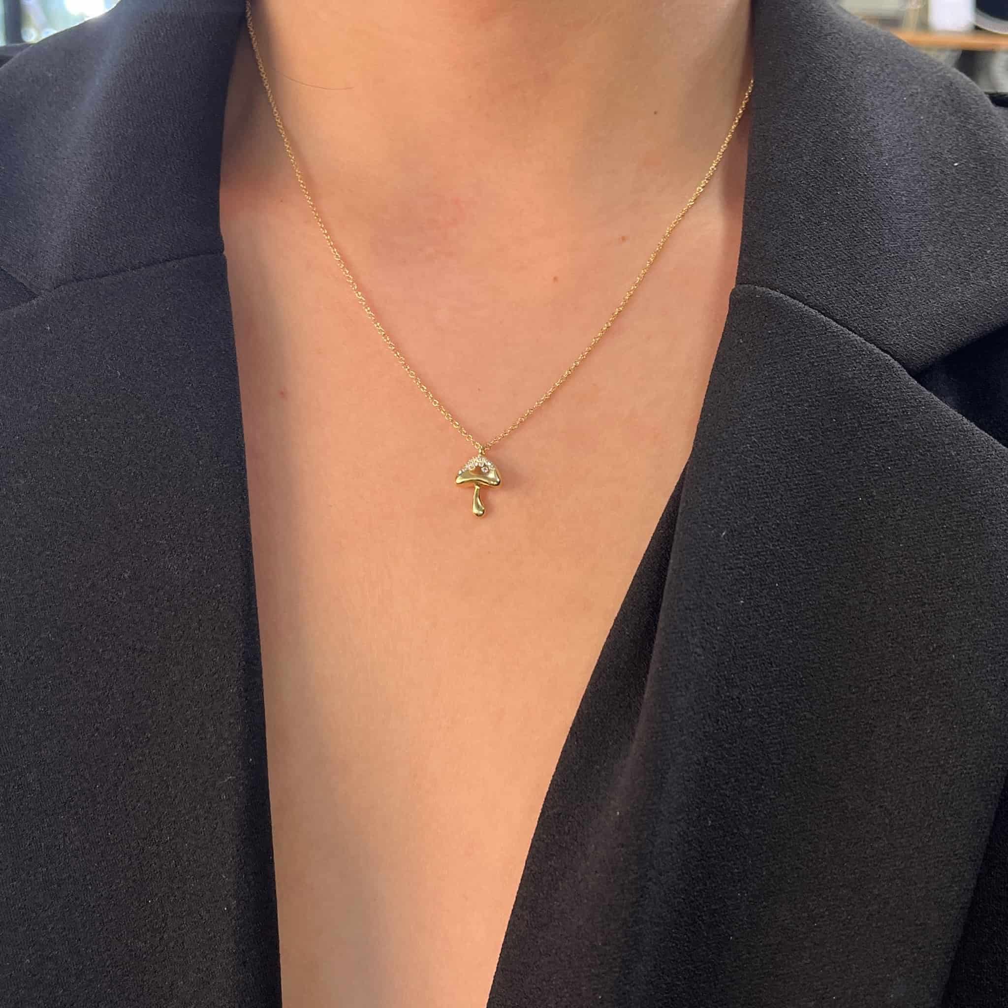 A person wearing a black blazer with a deep neckline showcases the Mini Diamond Mushroom Necklace from our fine jewelry collection, featuring a small mushroom pendant that contrasts beautifully against their skin and fabric.