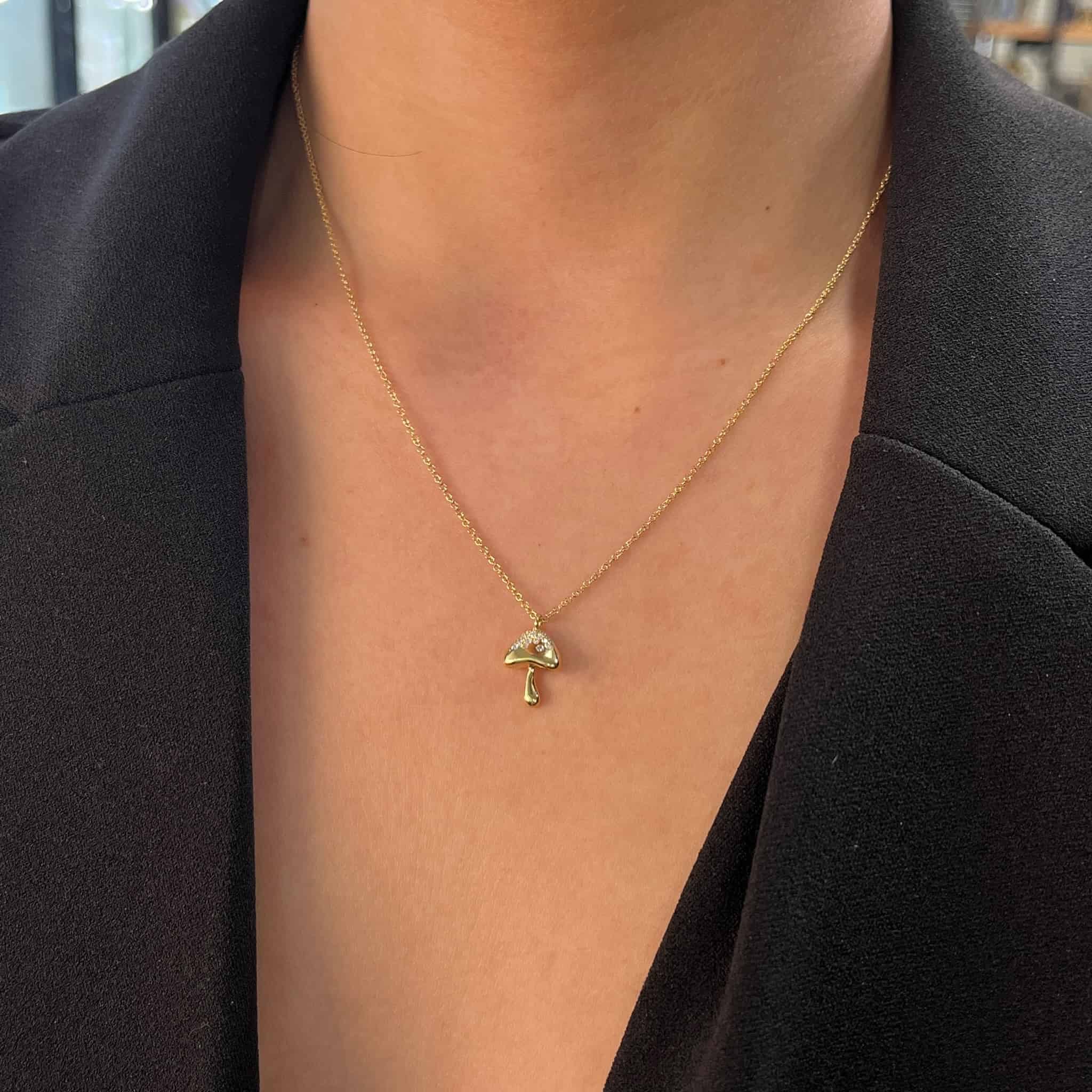 A person dons the Mini Diamond Mushroom Necklace, its custom mushroom-shaped pendant gleaming in gold against their black blazer, with a softly blurred background.