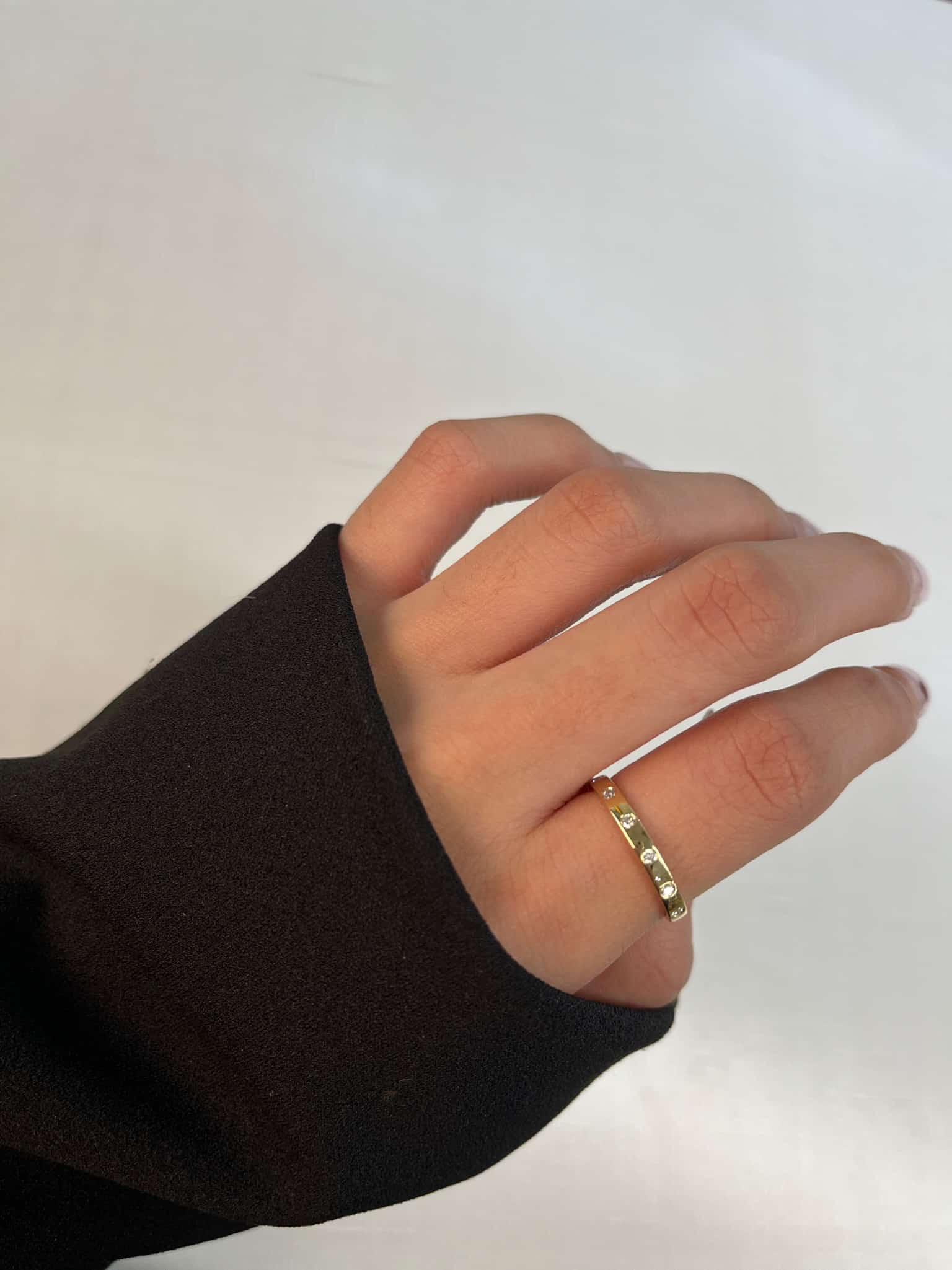 A hand elegantly draped in a black sleeve showcases the Diamond Avery Ring with its simple gold design on the ring finger. This fine piece of jewelry stands out against the light background, capturing attention and sparking curiosity.