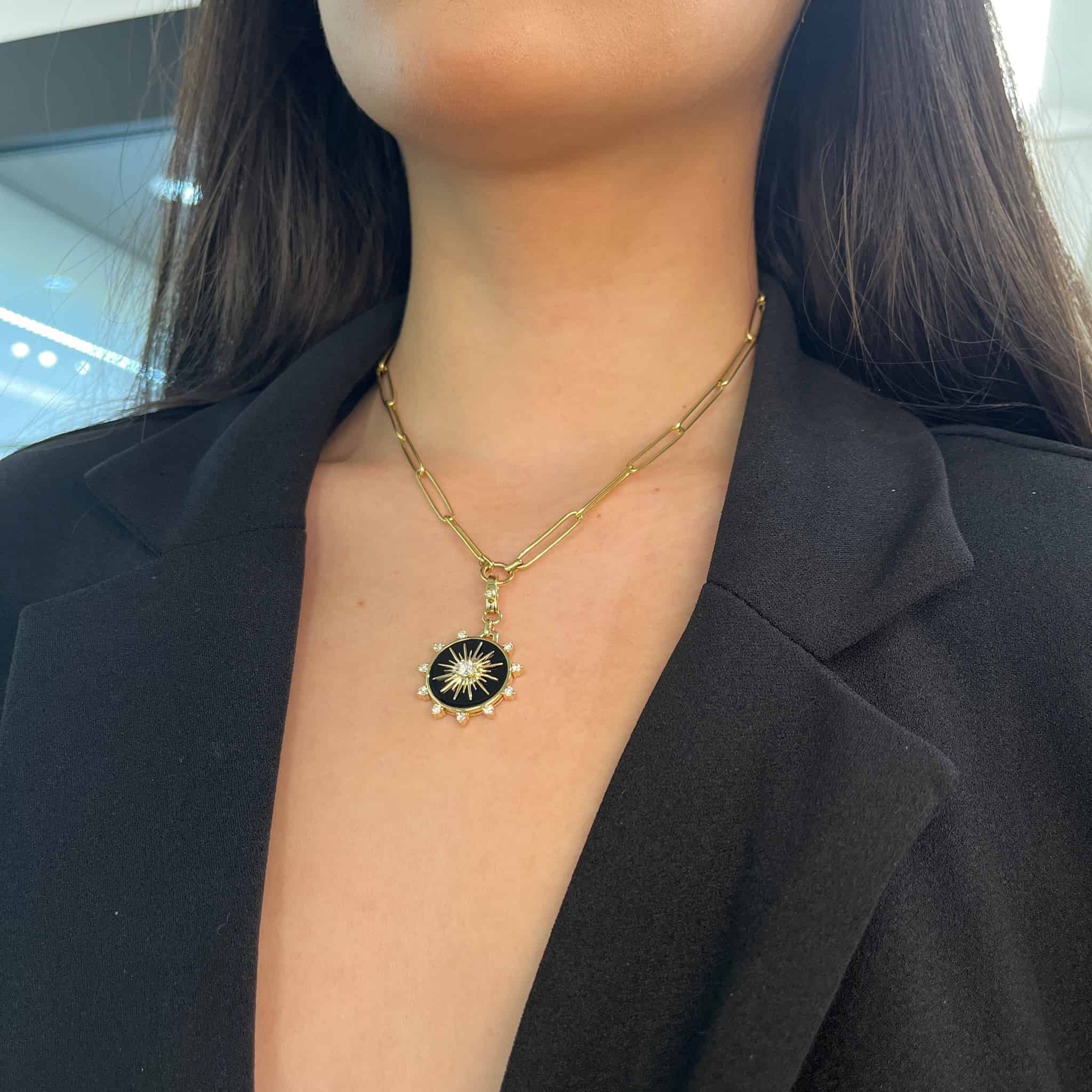 A person in a black blazer wears a gold chain necklace featuring the Diamond & Onyx Bentley Charm by Janet Heller Fine Jewelry, with a round starburst pendant against a softly out-of-focus background, creating an air of mystery.