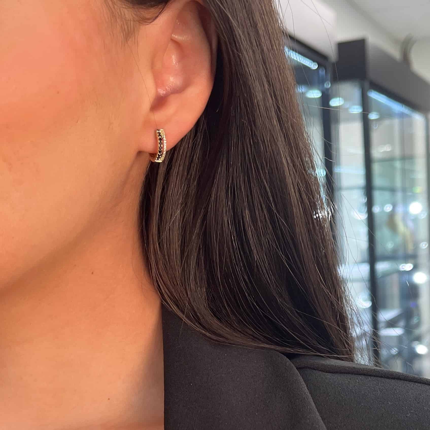 A close-up of a person with long dark hair wearing Double Row Black Diamond Huggies, set against a blurred backdrop of glass display cases. The small gold hoops have fine details and complement their black top perfectly.