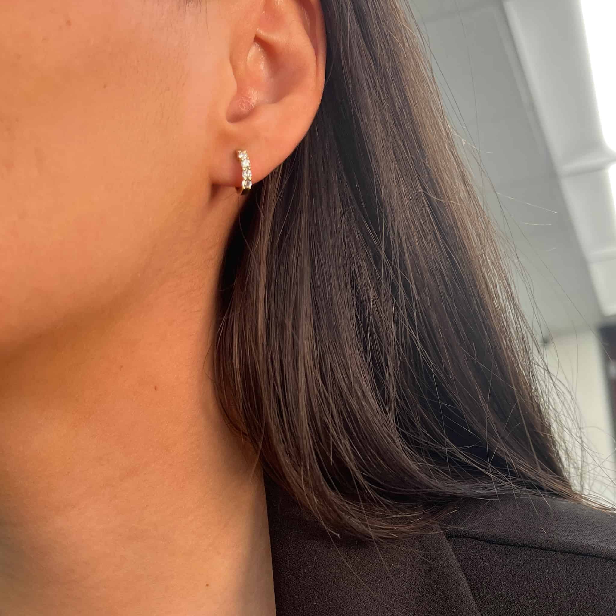 Close-up of a persons ear adorned with Diamond Dixie Huggies, small hoop earrings encrusted with tiny gemstones. The person has long, straight brown hair and wears a dark blazer. A softly blurred indoor background adds depth to the stylish image.