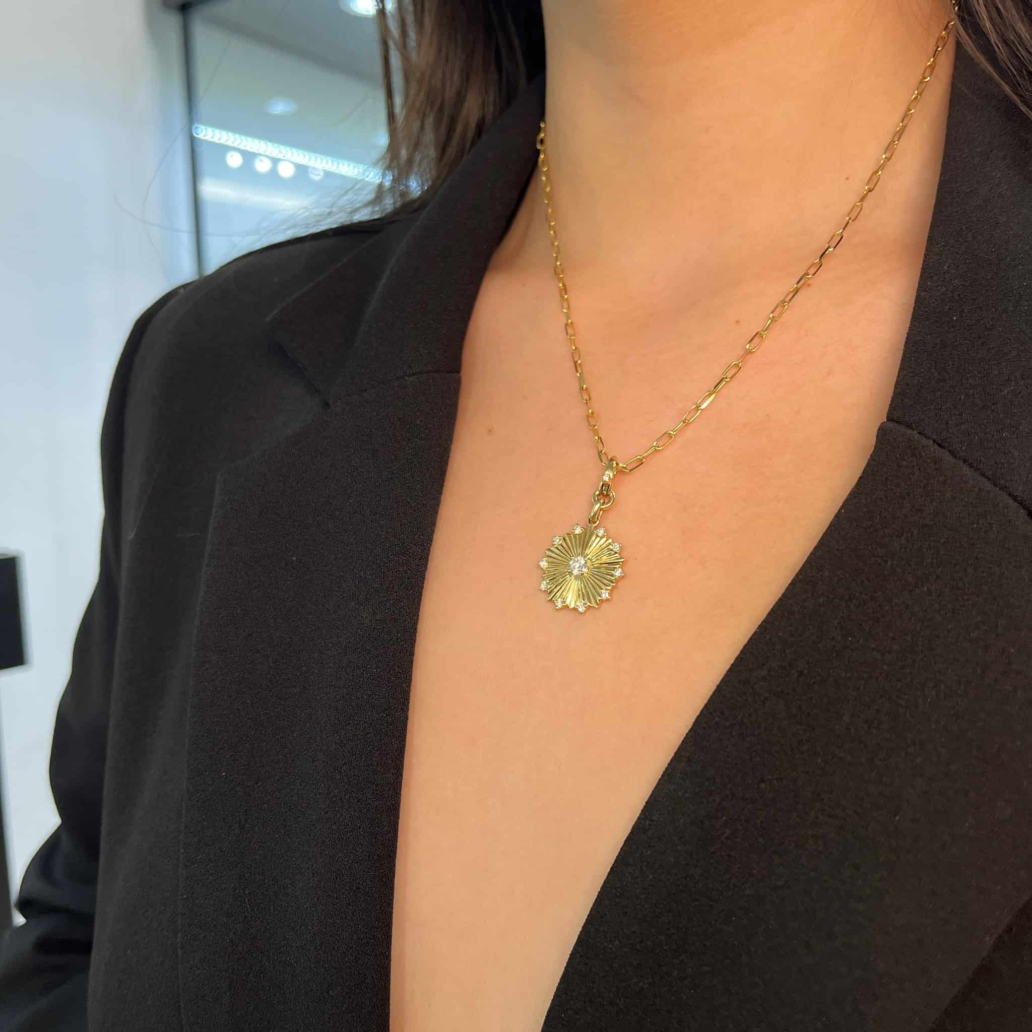 A woman in a black blazer displays the Diamond Disc Burst Charm by Janet Heller Fine Jewelry, highlighting its elegant clasp and intricate details against a subtly blurred backdrop.