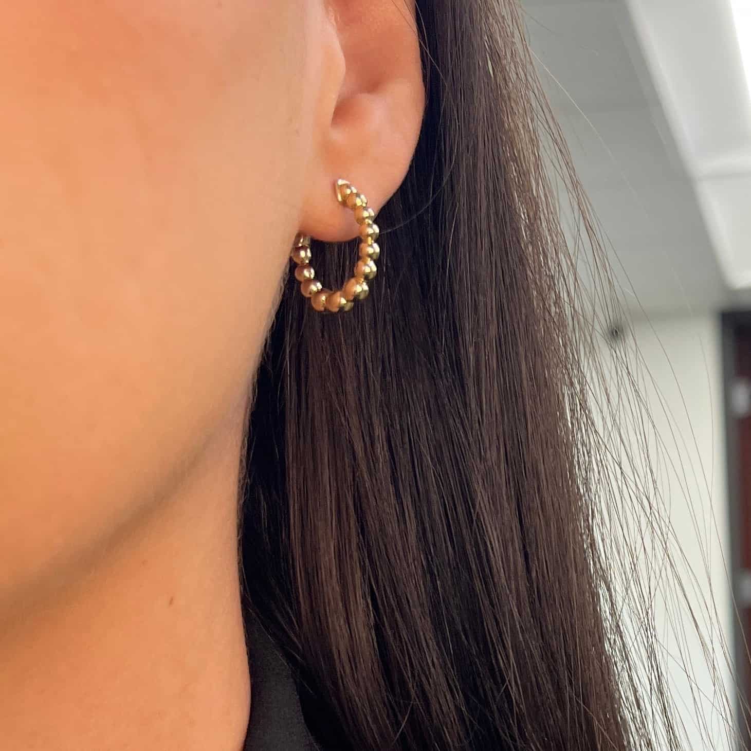 A close-up of a person's ear wearing the Janet Heller Fine Jewelry Mini Nora Hoops (SKU: EAR-1173-Y, $1058), featuring gold beaded detailing. The person has long dark hair and the background shows an indoor office setting. For custom jewelry or inquiries about this piece, please contact us.