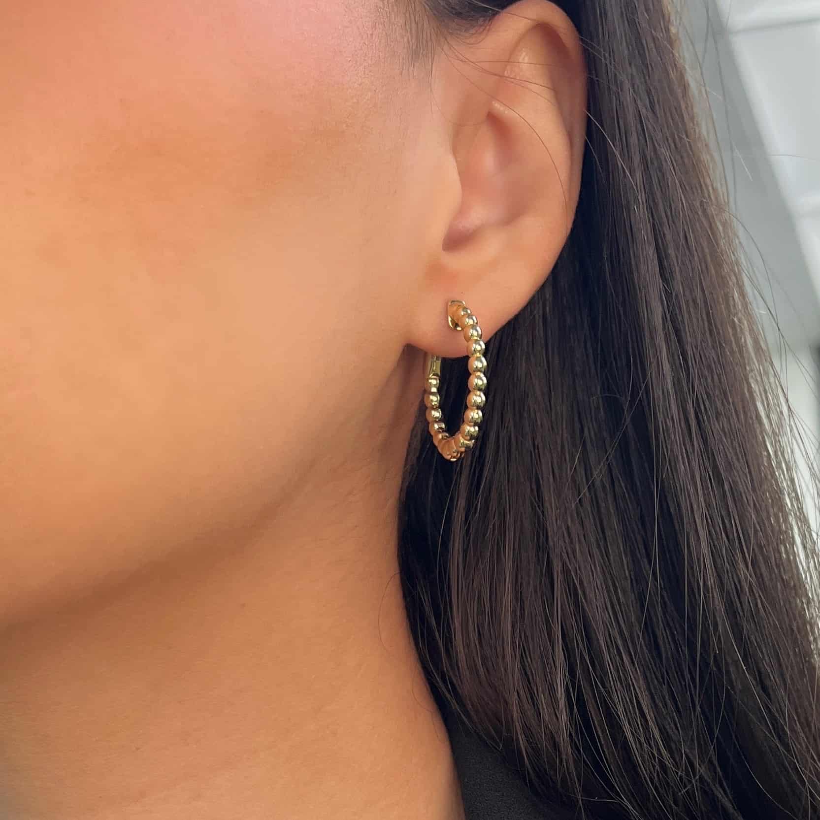 Close-up of a person’s ear wearing the Small Nora Hoops (EAR-1182-Y, 14k gold) from Janet Heller Fine Jewelry. The beaded gold earring is showcased against softly blurred background, as the model with long, straight dark brown hair and light skin poses elegantly. For custom jewelry or inquiries about the Small Nora Hoops, please contact us.