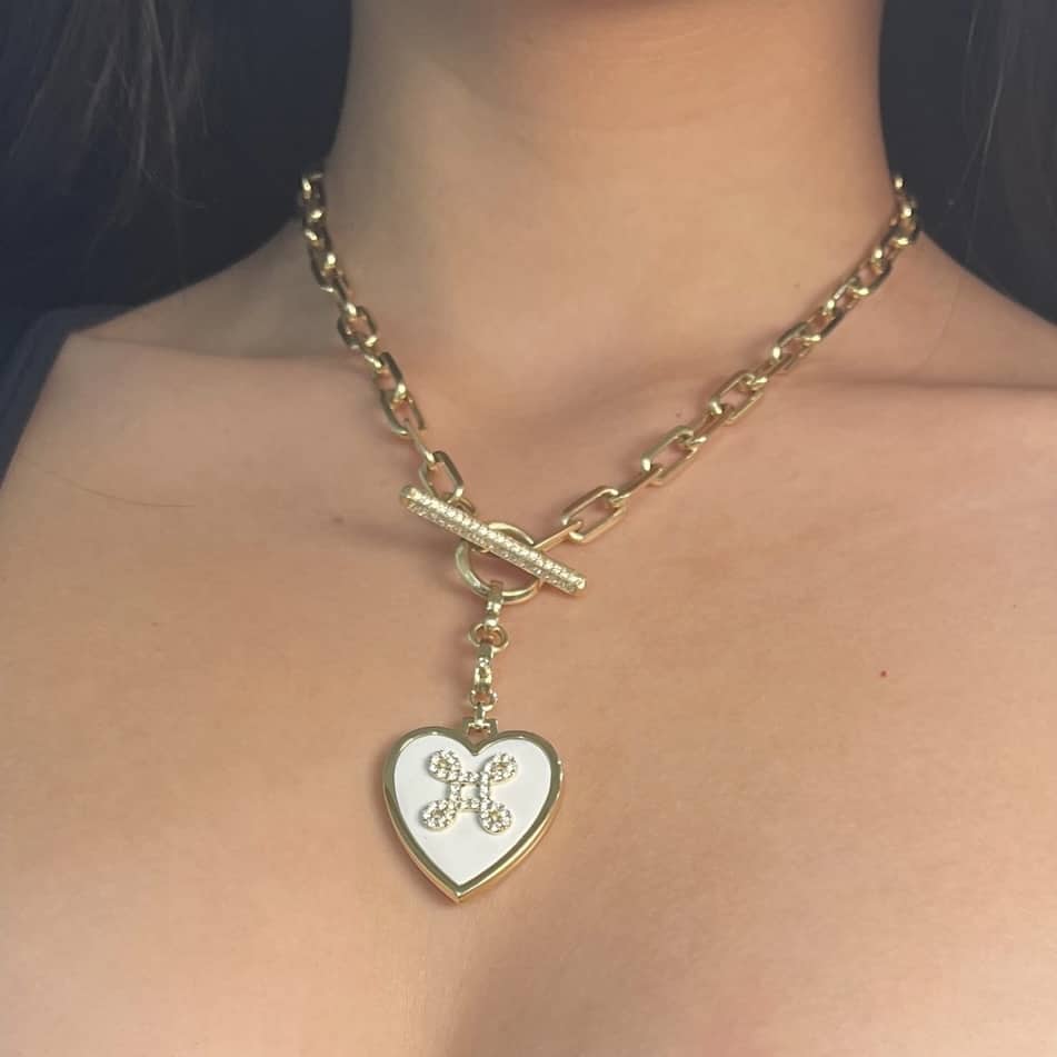 The White Heart Love Knot Charm Necklace from Janet Heller Fine Jewelry adorns a womans neck, boasting a floral heart-shaped pendant. With a toggle clasp at the front, it enchants with its white and gold design.