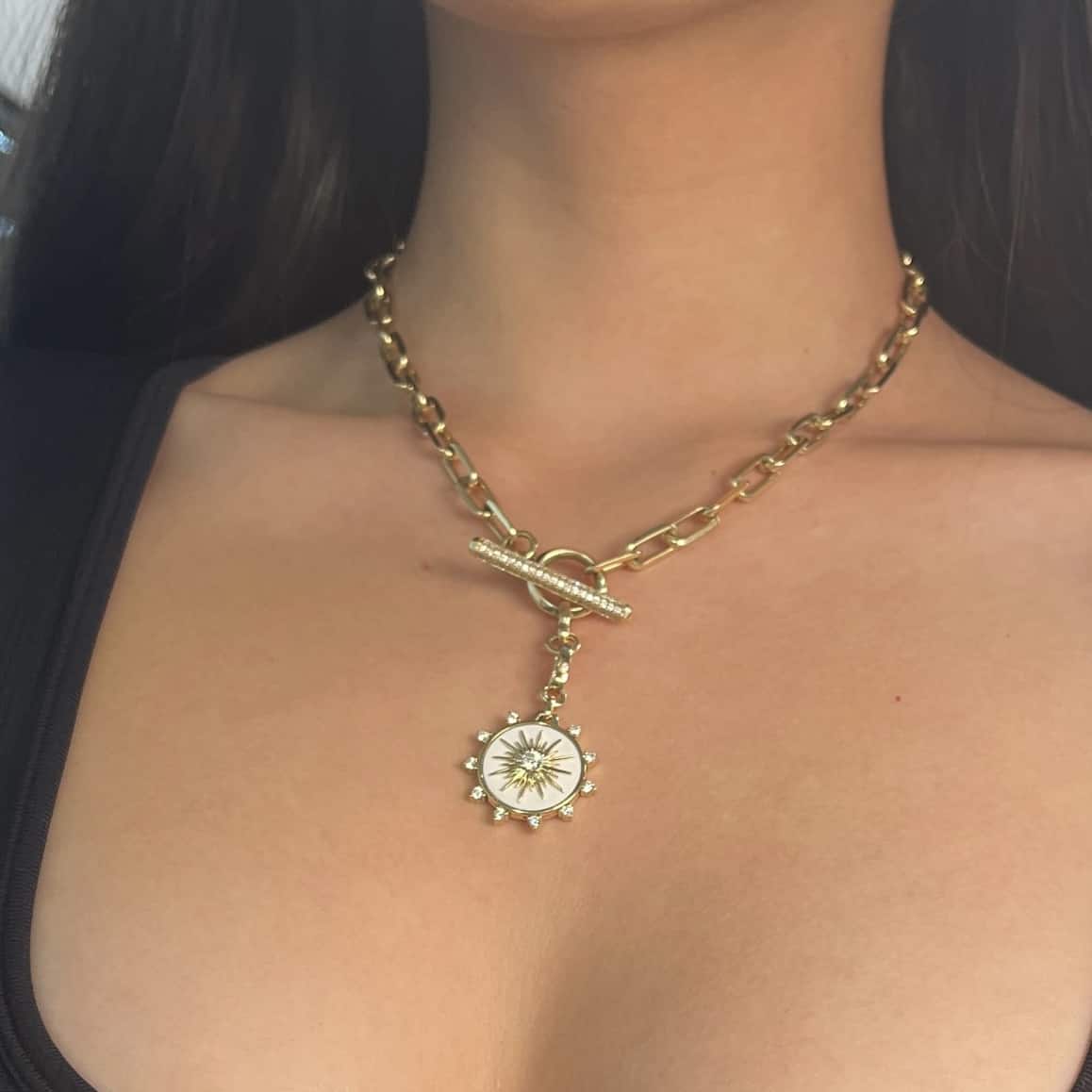 A woman elegantly pairs Janet Heller Fine Jewelrys gold chain necklace with a toggle clasp and sunburst pendant with a black top, her long hair flowing in the background. This piece can be endlessly customized with personal jewelry like the Diamond & Cacholong Bentley Charm.