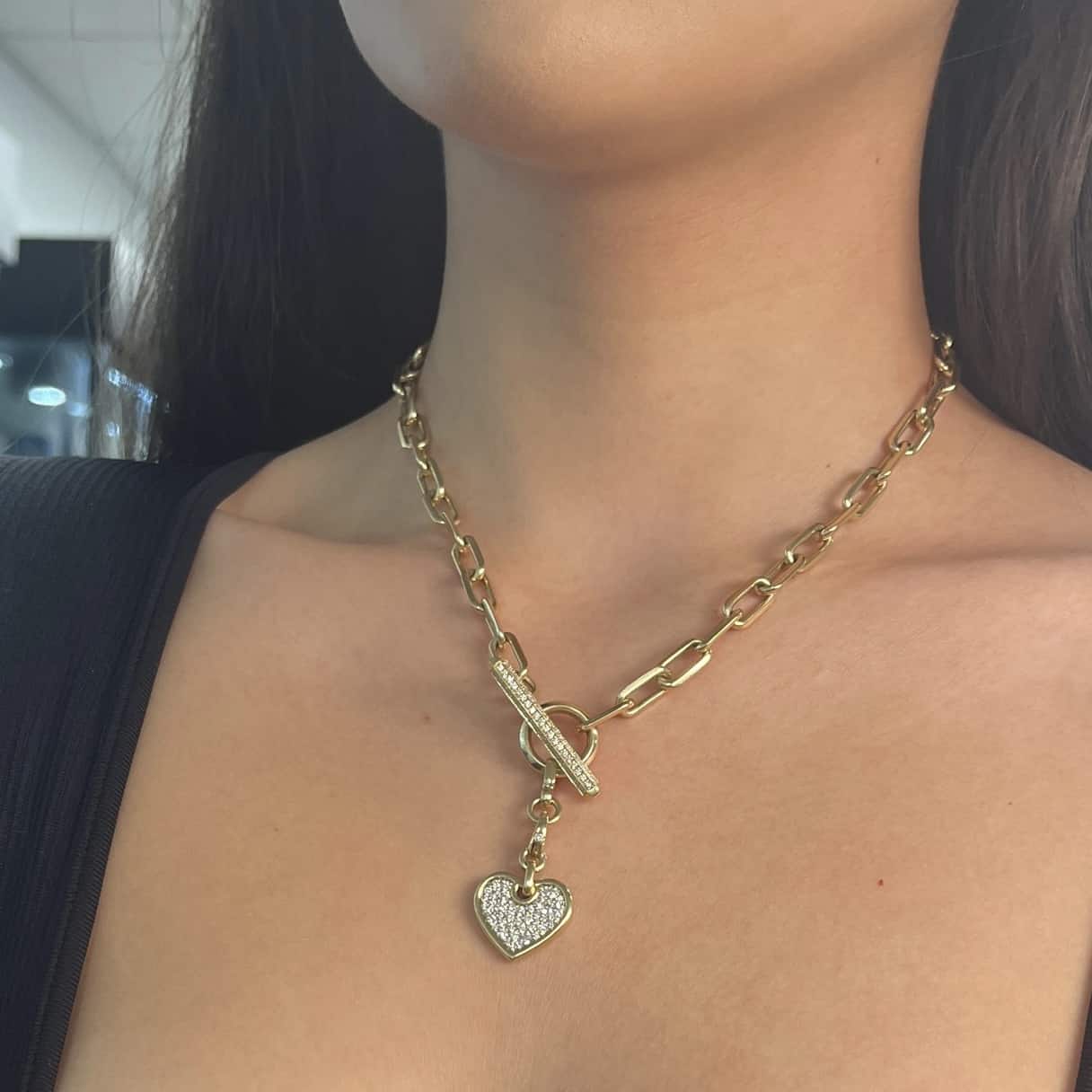 A person wears a Janet Heller Fine Jewelry gold chain necklace featuring a Medium Flat Diamond Heart Charm pendant. The elegant piece with a large toggle clasp stands out against a softly blurred background, highlighting its sparkle and sophistication.