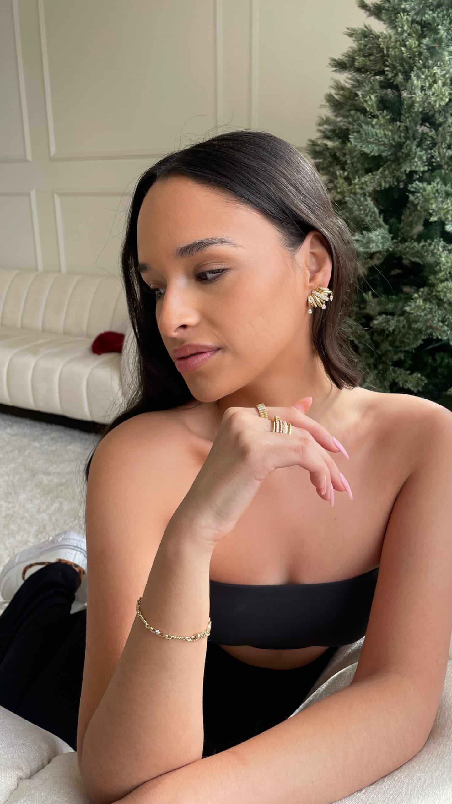 A woman with long dark hair sits on a light-colored couch, wearing a strapless black top and showcasing Janet Hellers Diamond Cage Earrings. Her chin rests on her hand, with a decorated Christmas tree sparkling in the background, enhancing the scenes warm ambiance.