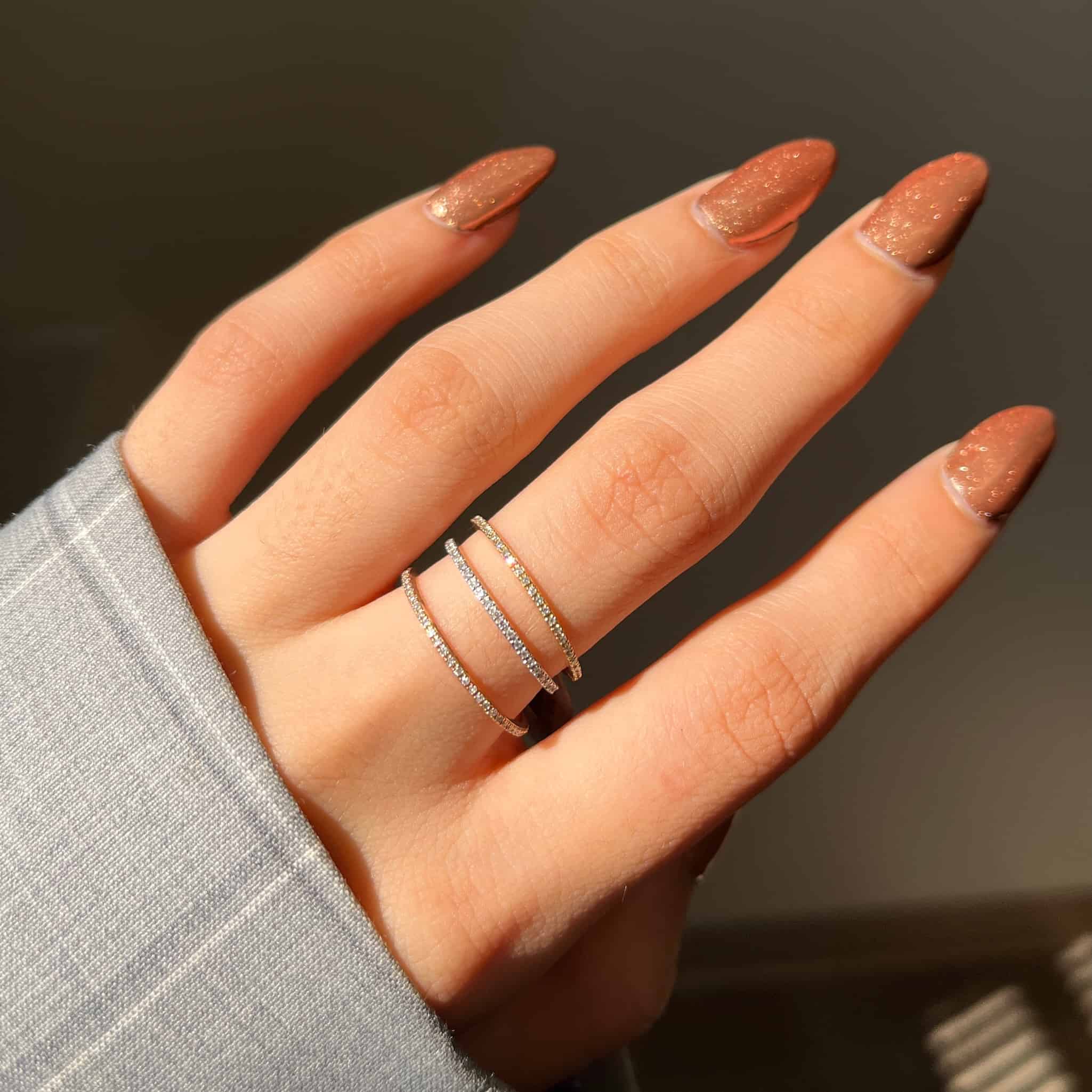 A hand with peach-colored glittery nails flaunts two Micro Pavé Eternity Rings on the middle finger, epitomizing classy jewelry elegance. Partially covered by a light gray plaid sleeve, the softly blurred background adds mystery. Preorder now for timeless style.