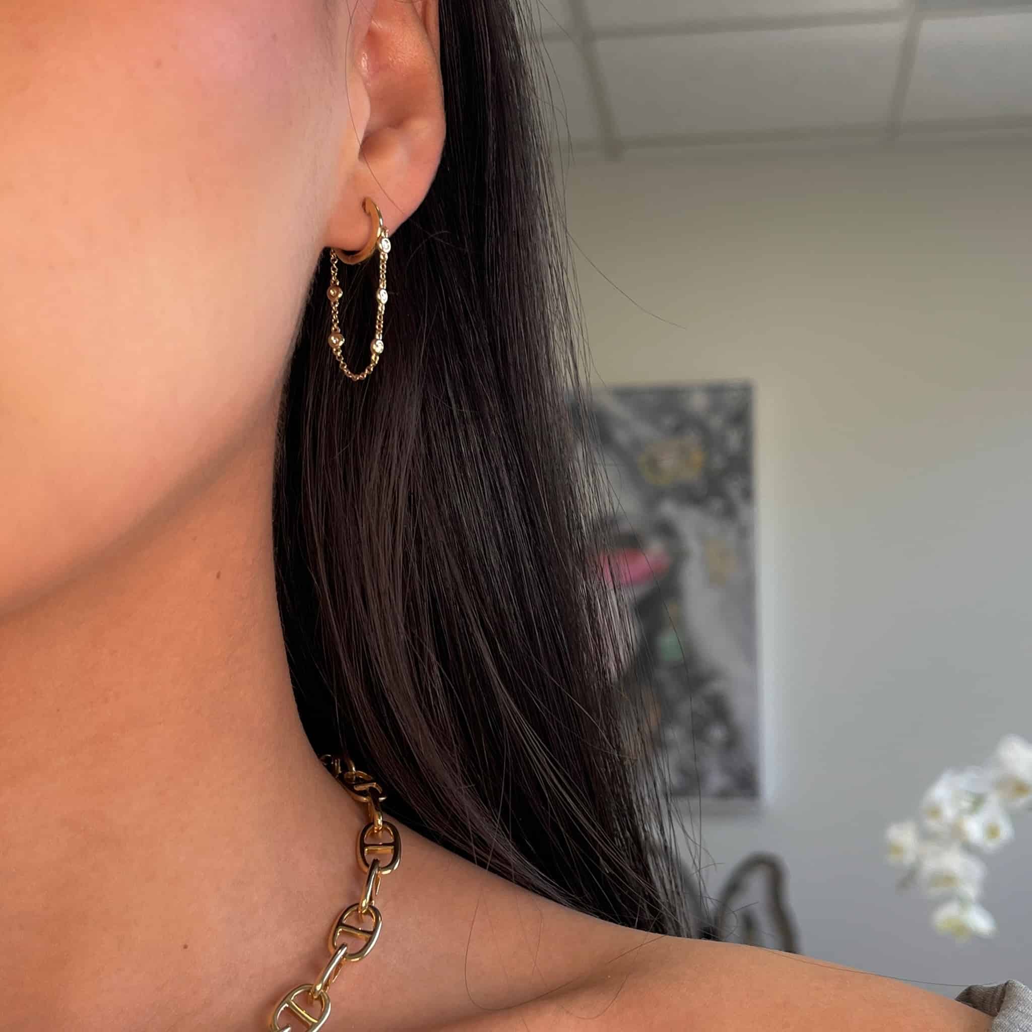 A close-up features a person wearing Diamond Chain Quinn Earrings, paired with a chunky gold necklace. The intricate customizations are highlighted against a softly blurred background with abstract art and a white flower, subtly emphasizing each product detail.