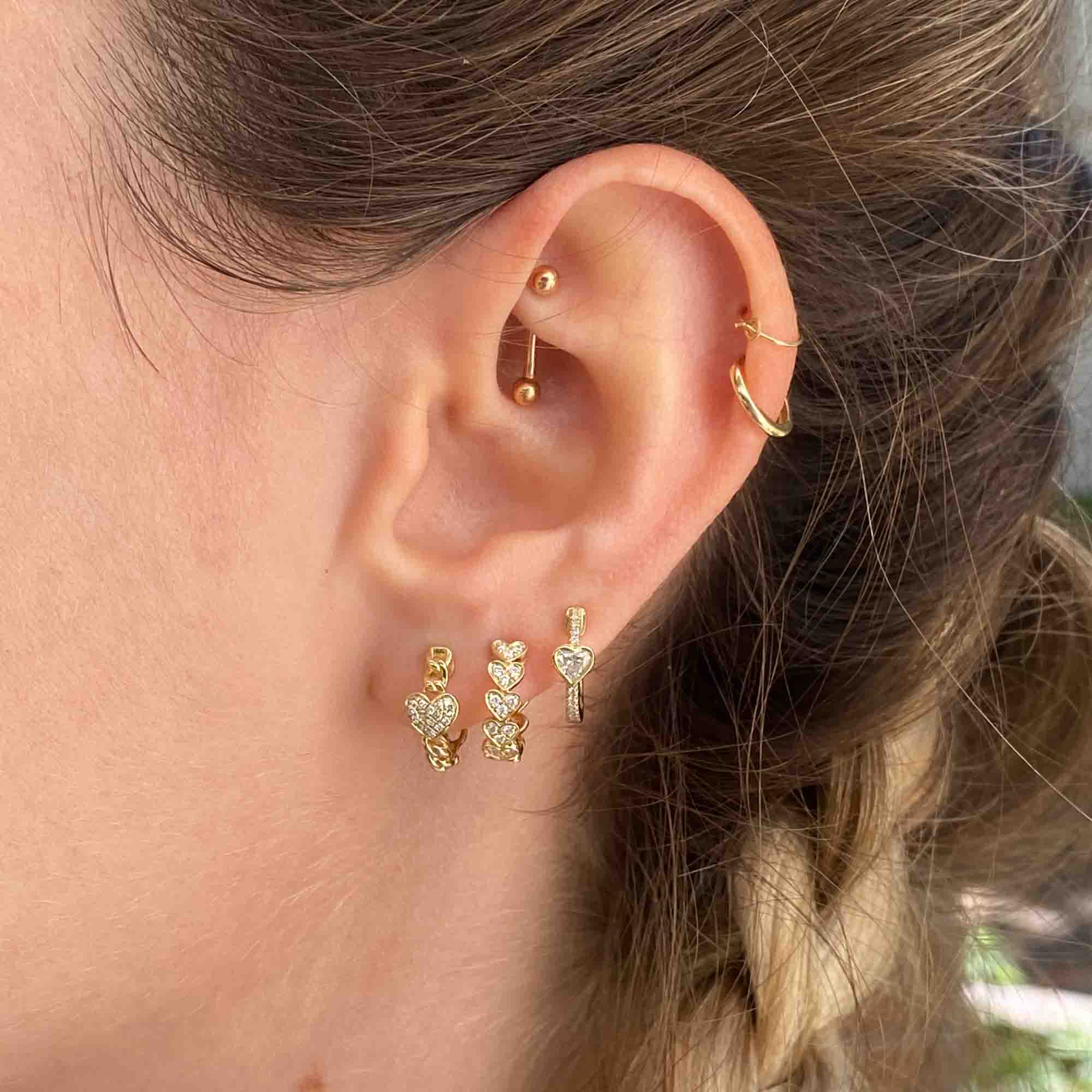 A close-up of a womans ear displays fine jewelry with multiple earrings: a gold hoop, barbell piercing, and Diamond Amore Huggies. Her hair is styled in a braid, enhancing the elegance.