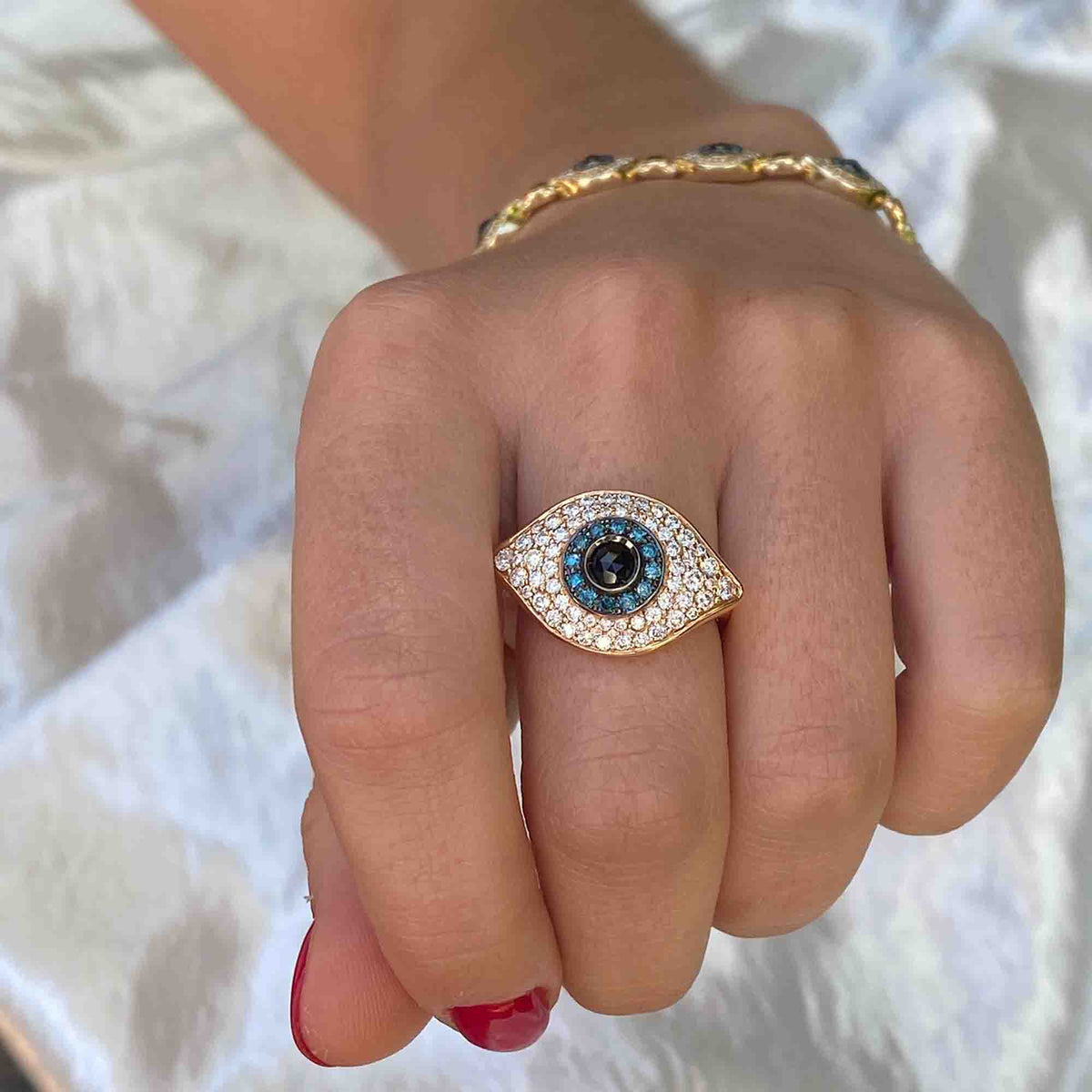 A hand with red nail polish wears the Large Diamond Evil Eye Ring—a gold ring shaped like an eye and adorned with white and blue stones—paired with a matching gold bracelet. For custom ring sizes or to preorder this unique piece, please contact us.
