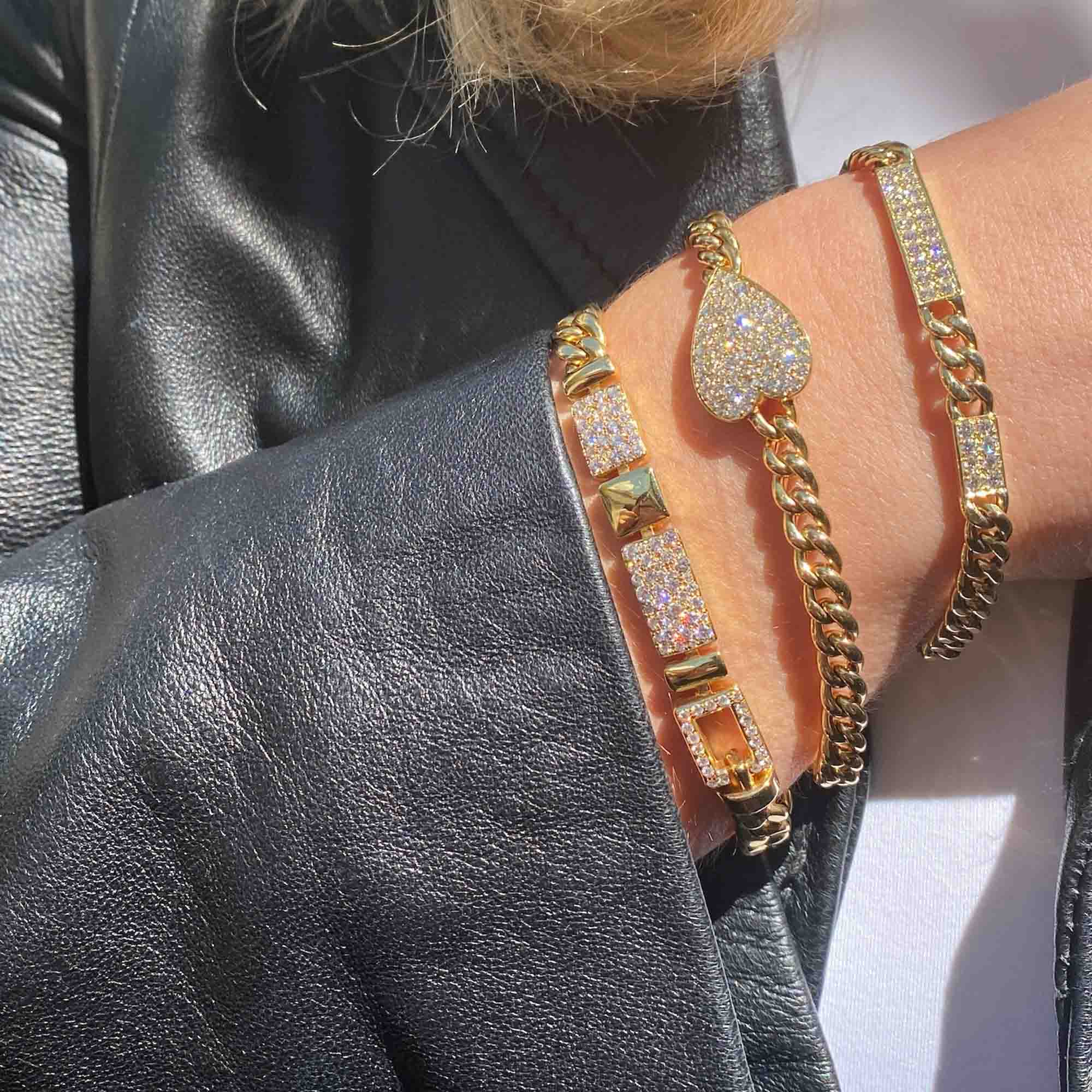 A person in a black leather jacket showcases three stunning jewelry pieces on their wrist, including a gold Cuban Link Diamond Bars Bracelet with sparkling stones and customizations like heart designs, chain links, and square accents.