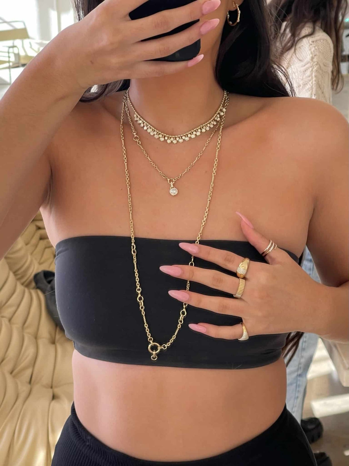 A woman wearing a black tube top showcases the Diamond Dangle Toilet Chain Necklace, styled with layered gold rings and long pink nails—elegant pieces that reflect the artistry of Janet Heller Fine Jewelry. She holds a phone partially covering her face, with a beige couch and another person in the background.
