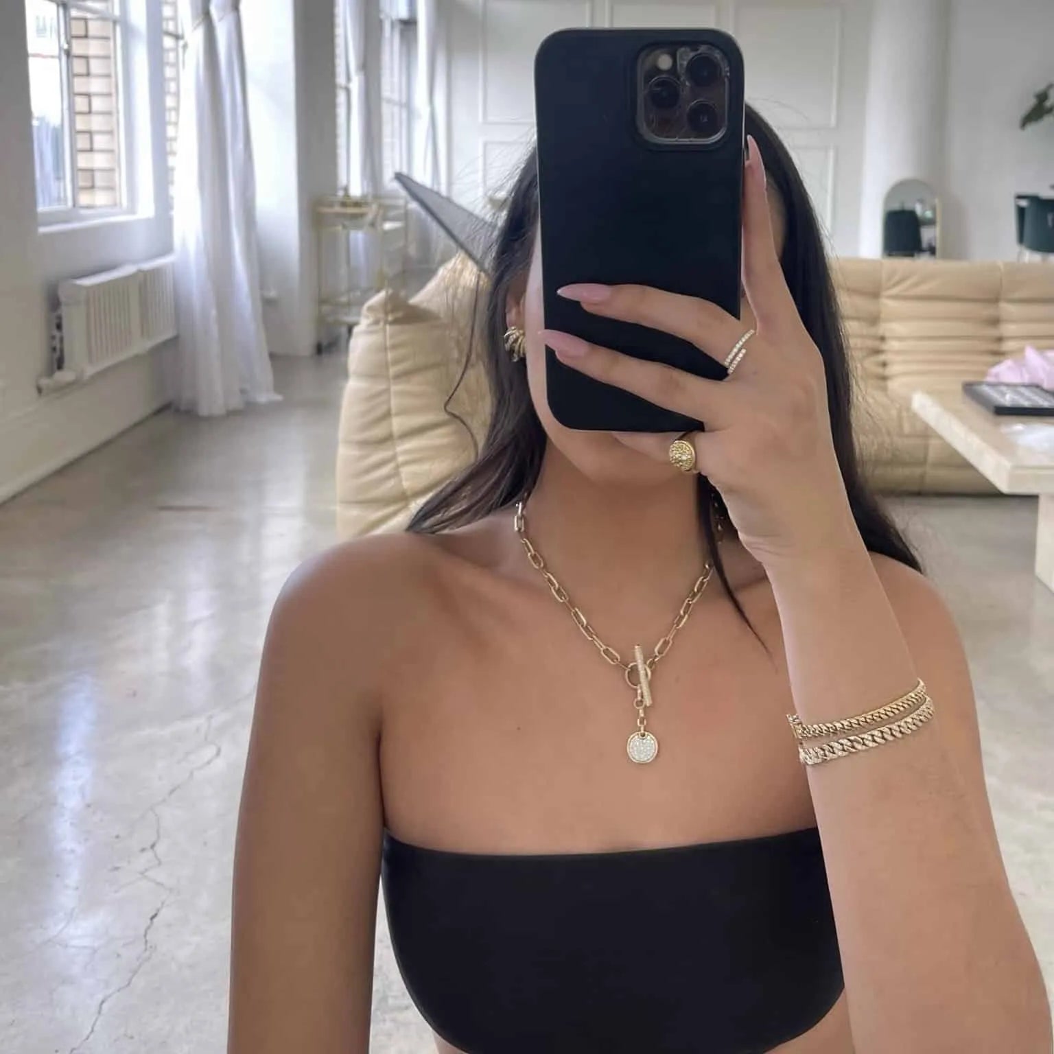 A person takes a mirror selfie wearing a black strapless top, hiding their face but showcasing long dark hair and Janet Hellers Diamond Toggle Link Milan Chain. The rooms modern vibe is enhanced by a beige couch and large windows.