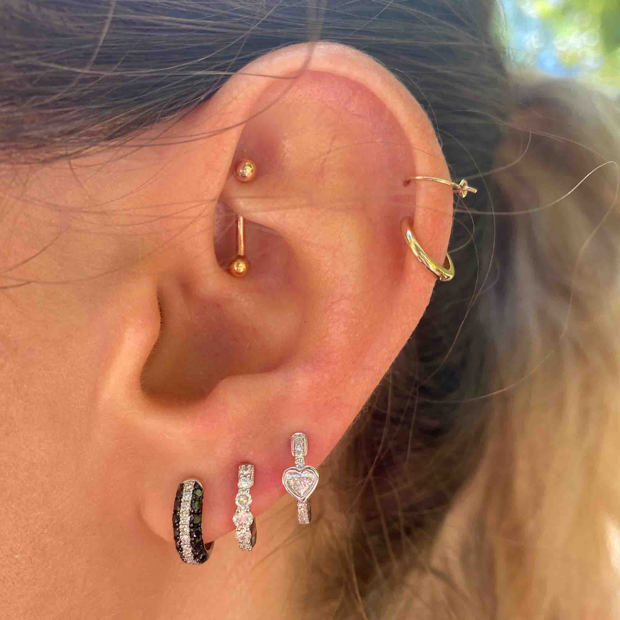 A close-up of an ear with multiple piercings, featuring three stone-adorned earrings, a heart-shaped piece, a gold hoop at the top, and two small gold studs in the inner cartilage—an elegant showcase of Janet Heller’s custom jewelry design highlighted by the Black & White Diamond Huggies ($725).